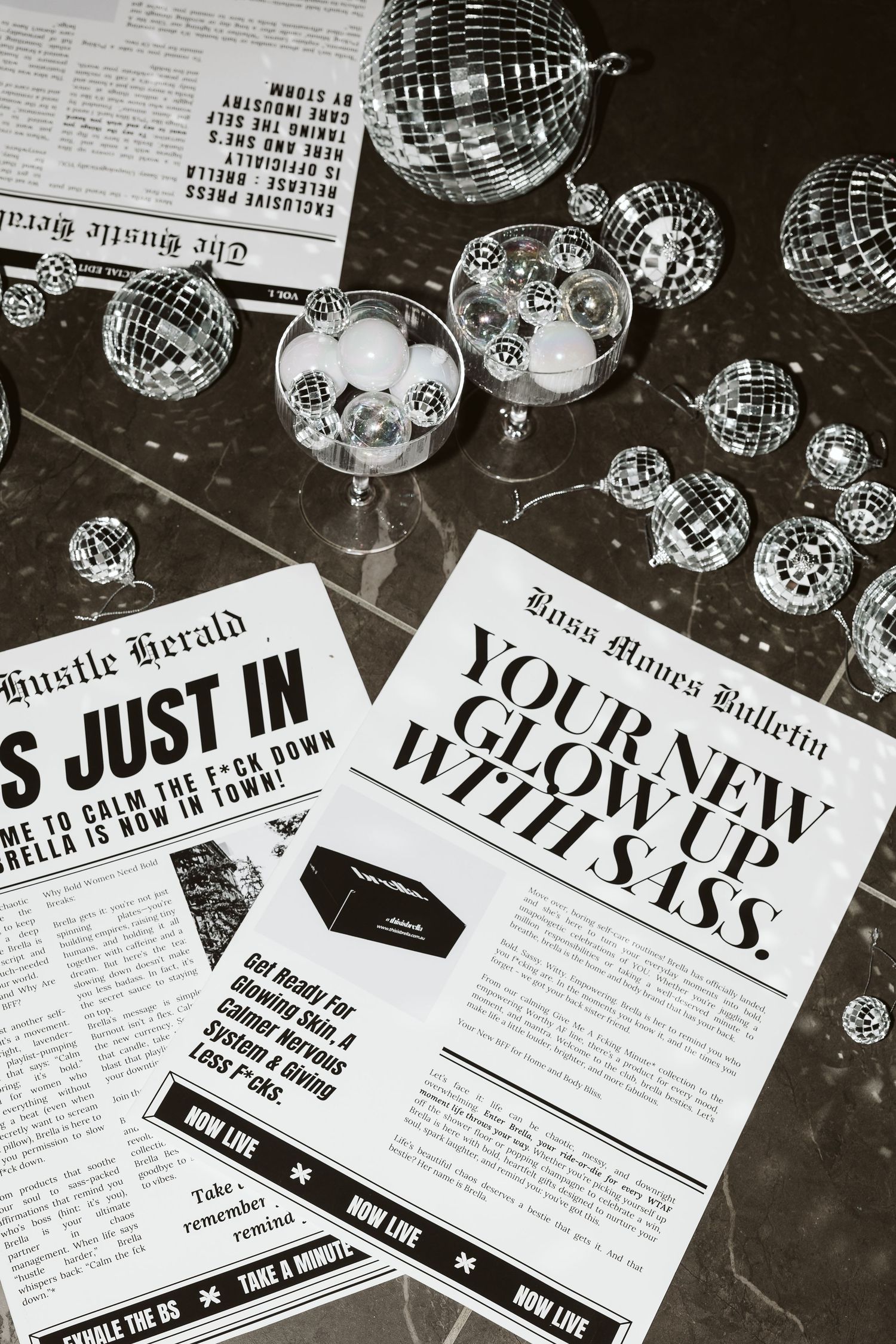 Black and white flat lay of Brella brand newspapers and disco balls on marble surface.