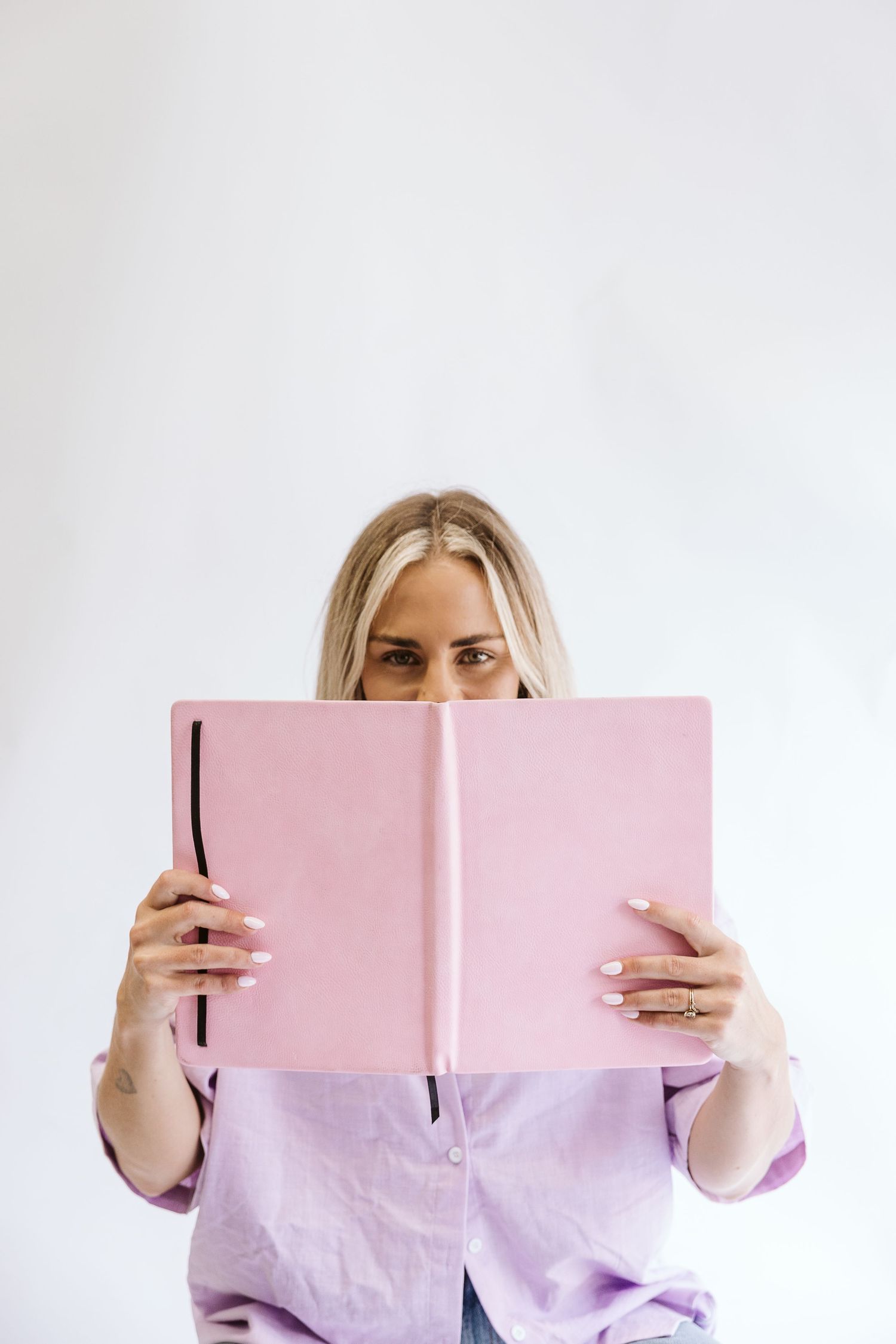 Blonde woman hiding face behind open pink notebook, peering over top with white manicured nails.