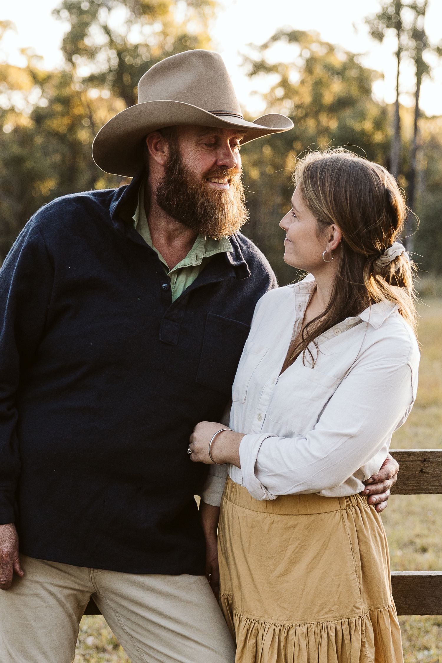 A bearded man in a cowboy hat and a woman in a white blouse share a warm moment outdoors at golden hour.