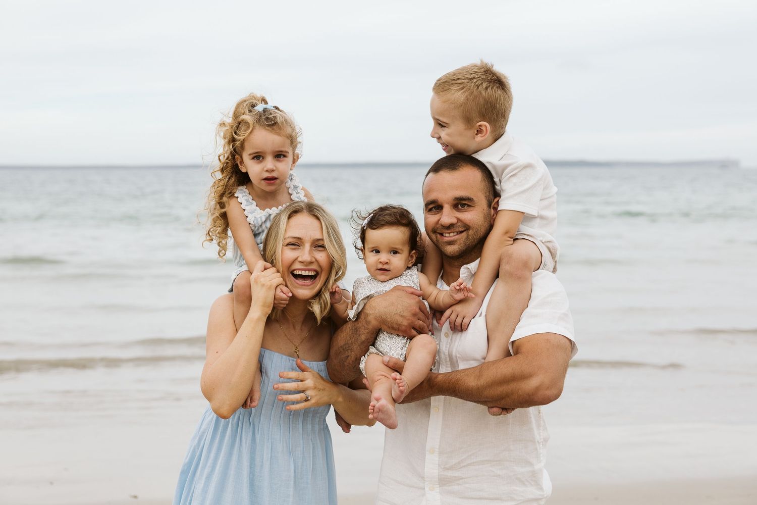 Happy family of five posing on a beach, parents holding three young children, smiling with ocean waves in the background.