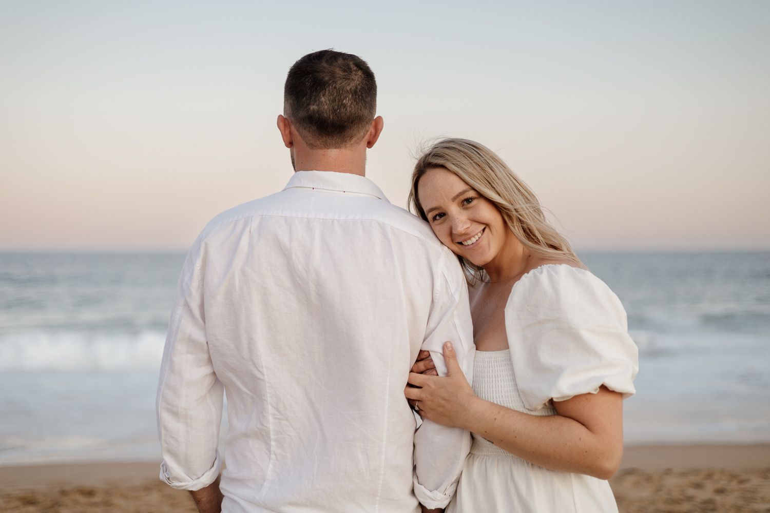 Blonde woman in white dress smiling over her shoulder while embracing her partner on a beach at dusk.