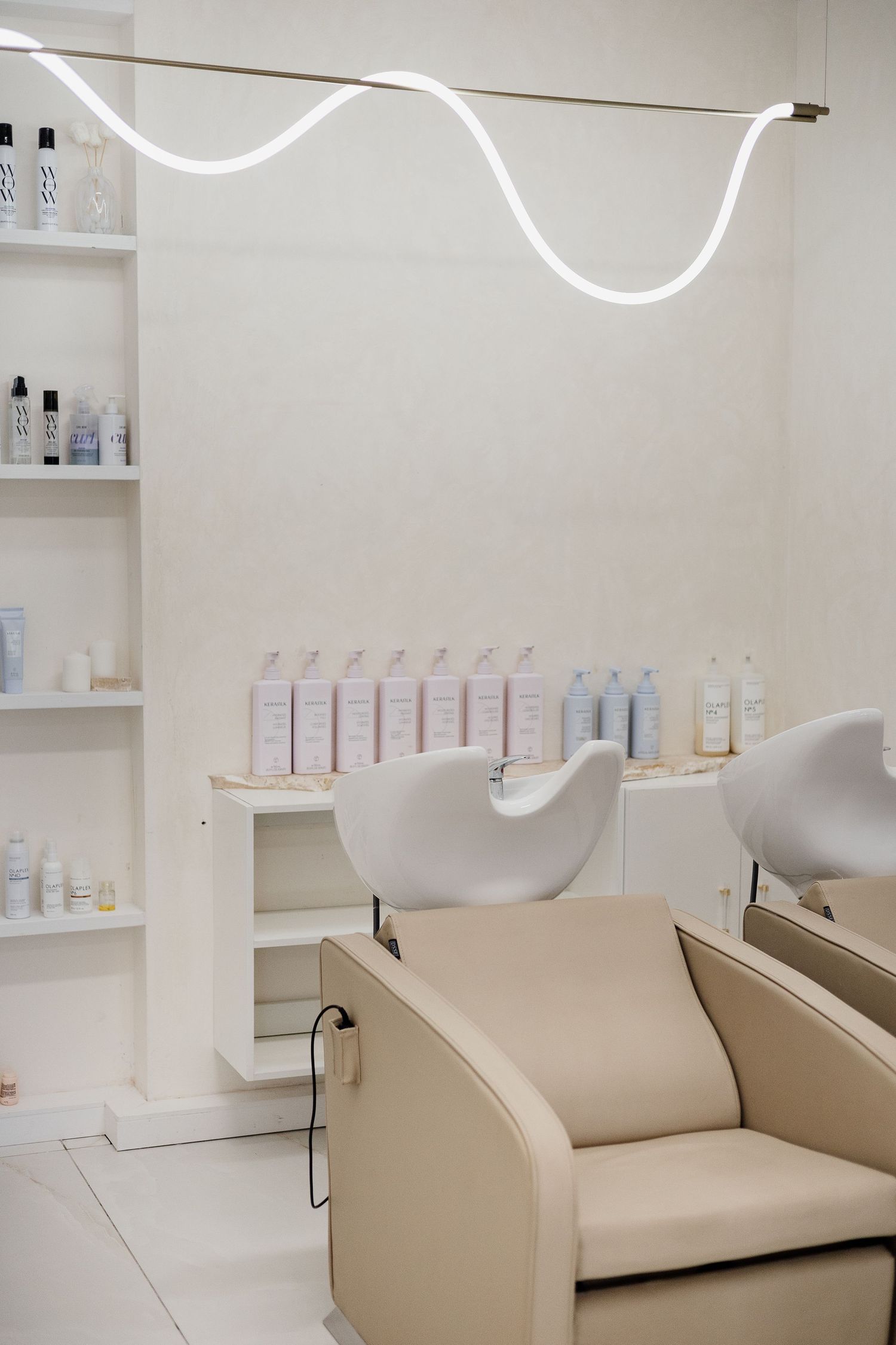 Luxury hair salon wash area with beige shampoo chairs, wavy neon light, and product shelving.