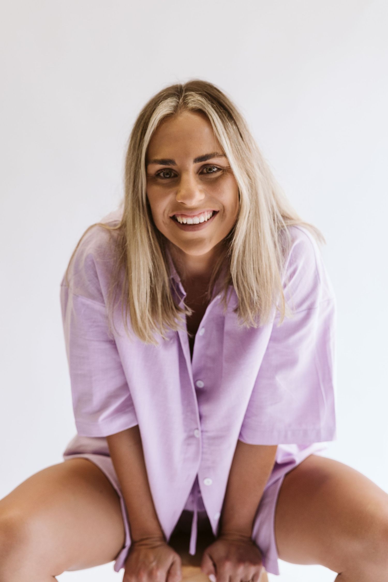 Laughing blonde woman in purple linen shirt leans forward on white background.