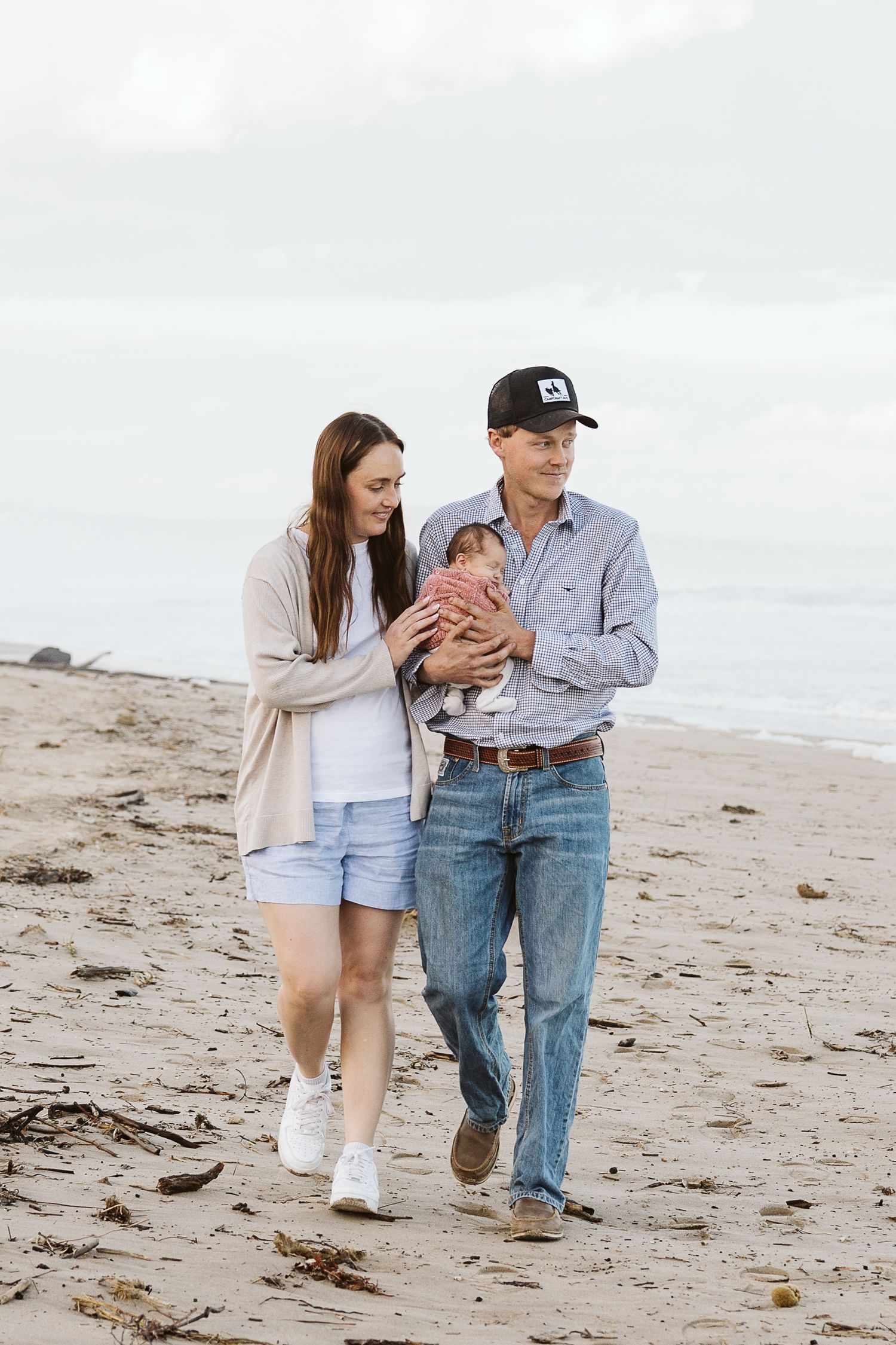 Happy young family walking on a sandy beach, father holding newborn baby in pink outfit.