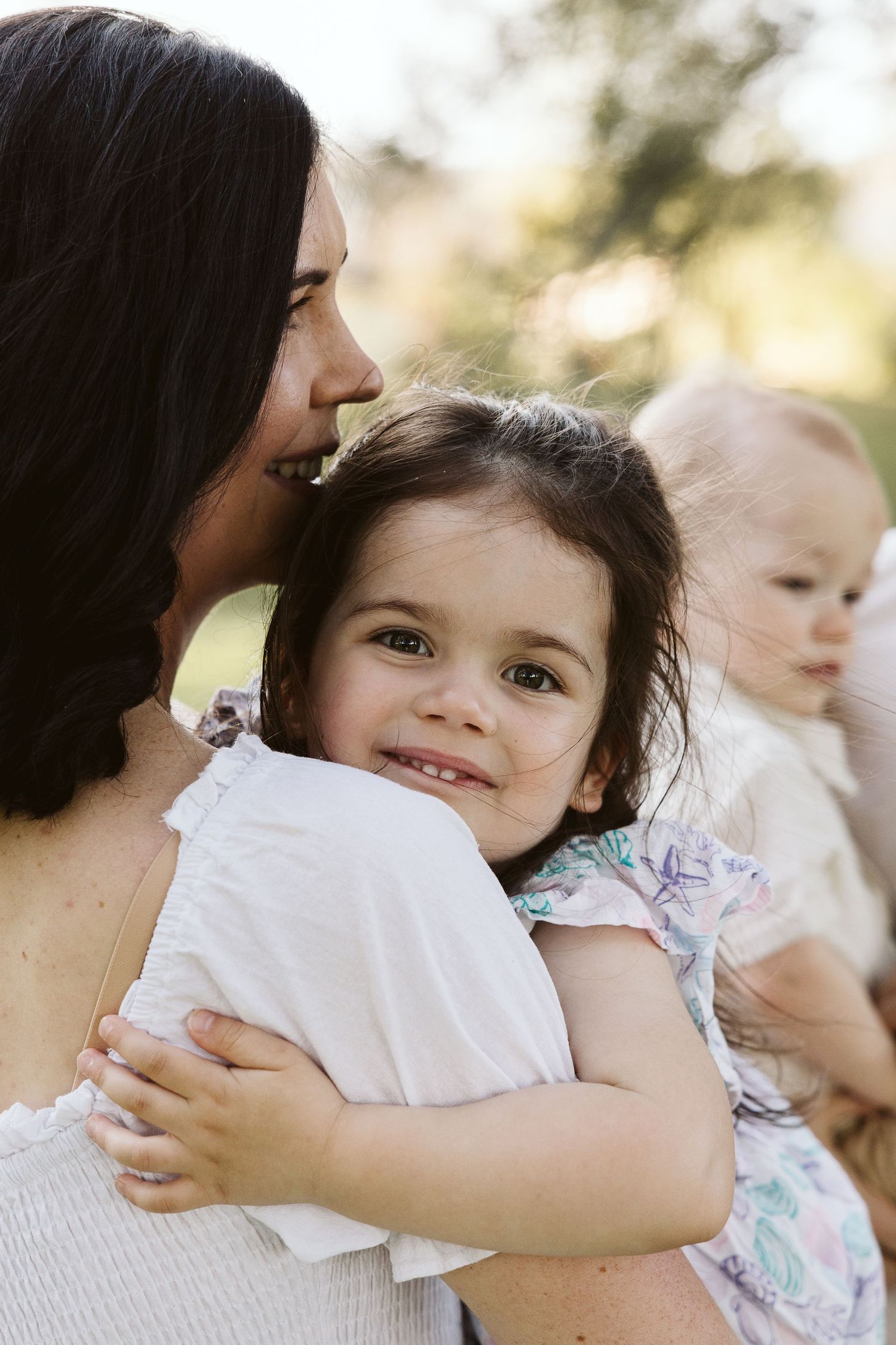 A mother kissing her smiling toddler daughter on the cheek while holding a baby outdoors in soft natural light.