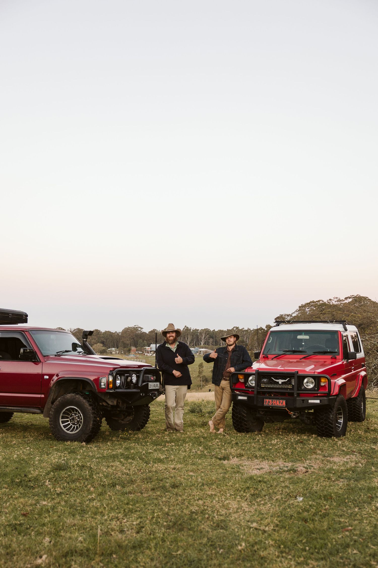 Two people stand between two red 4WD vehicles on an open grassy field at dusk in Australia.