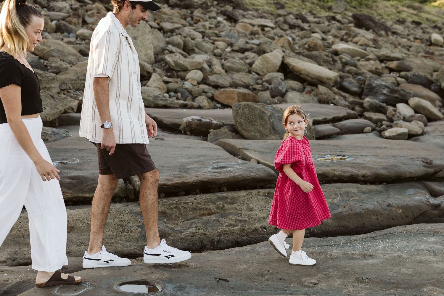 Family of three walking on rocky beach, young girl in pink dress leads parents dressed in white and black casual wear.