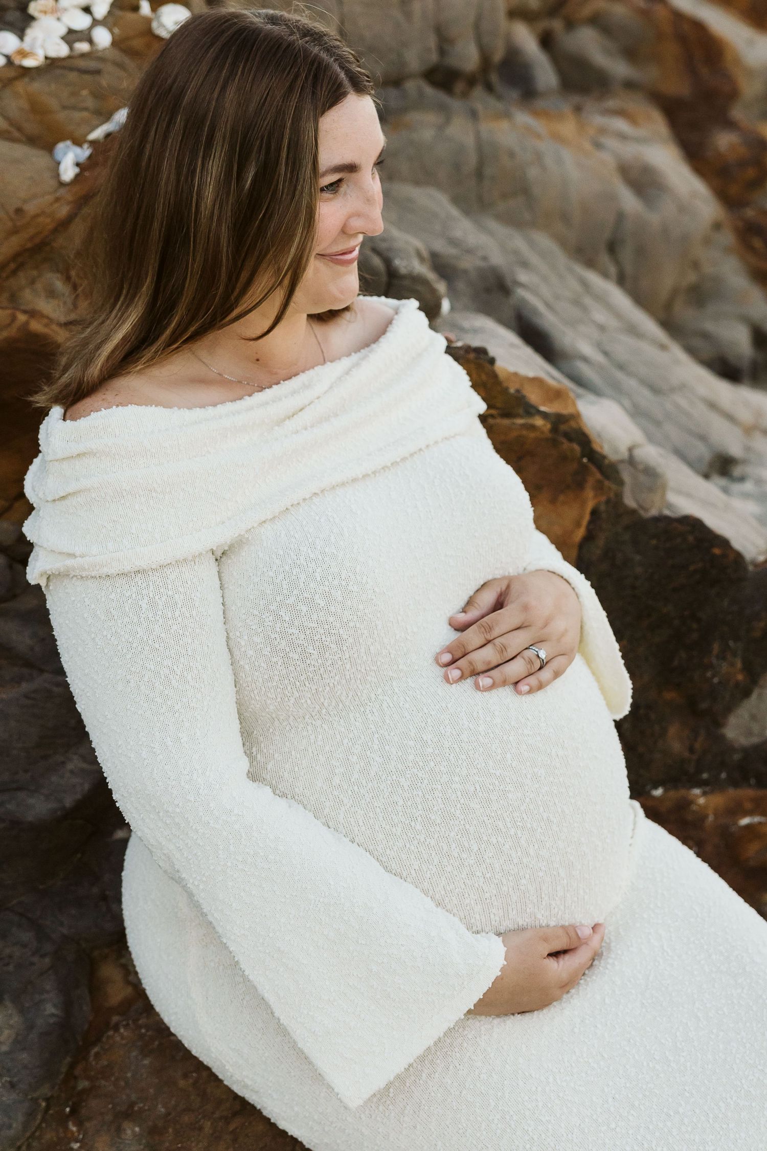 Smiling pregnant woman in white off-shoulder bouclé dress cradling her baby bump while sitting against rocky backdrop.