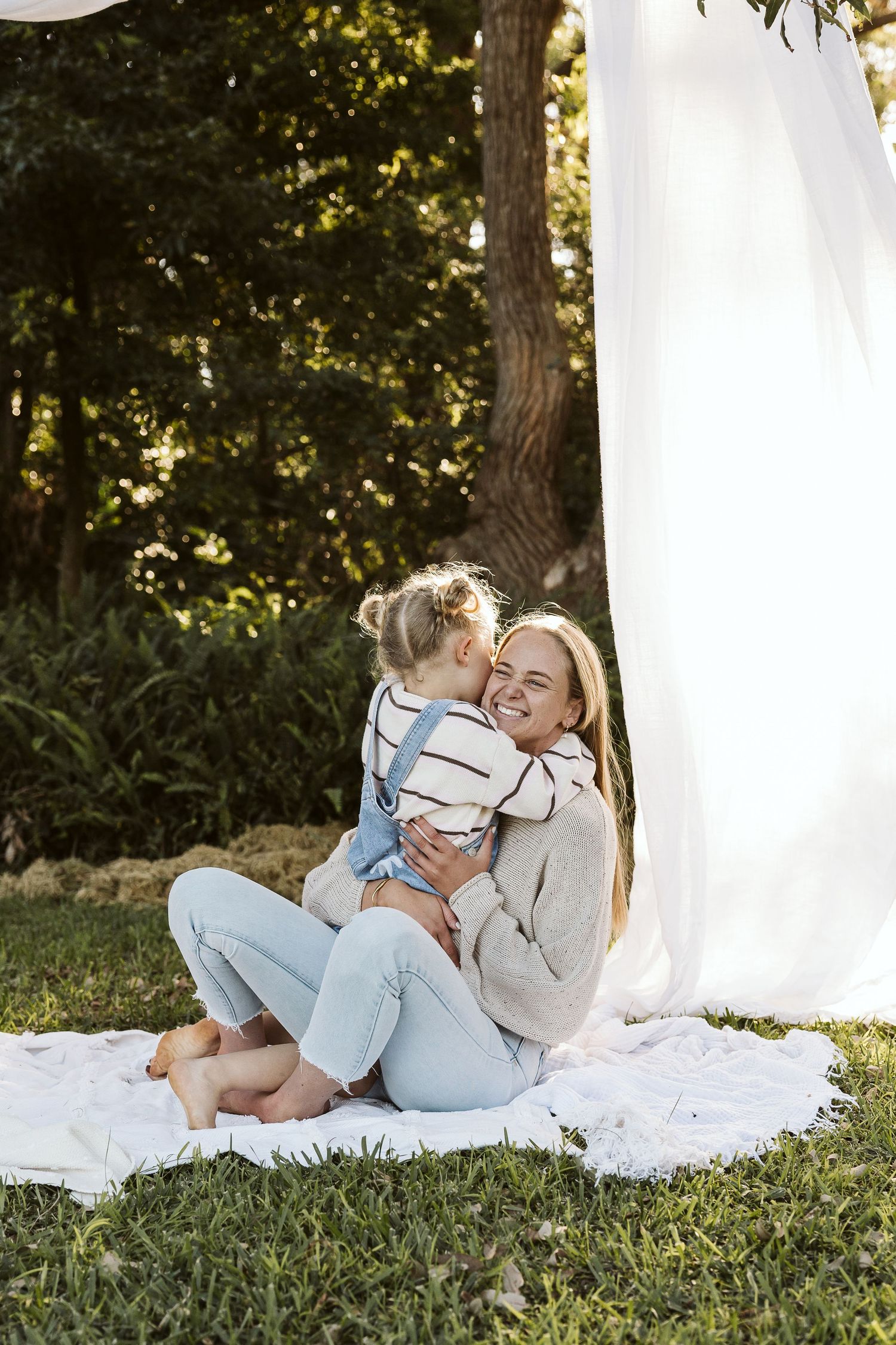 Mother hugging her young daughter on a blanket in a sunny outdoor garden setting. Motherhood photoshoot.