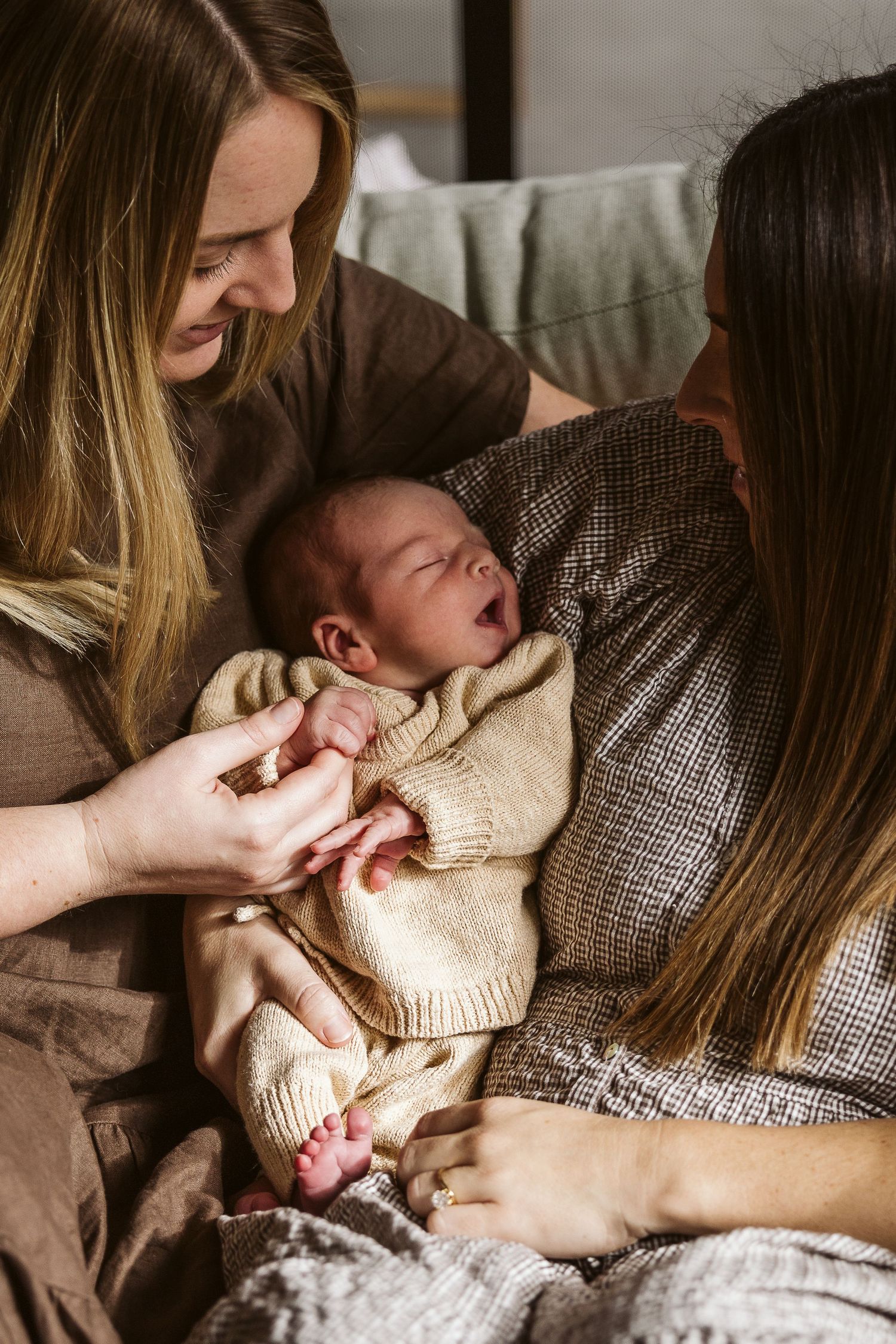 Two mothers smiling and bonding with their yawning newborn baby wrapped in cream knit sweater.