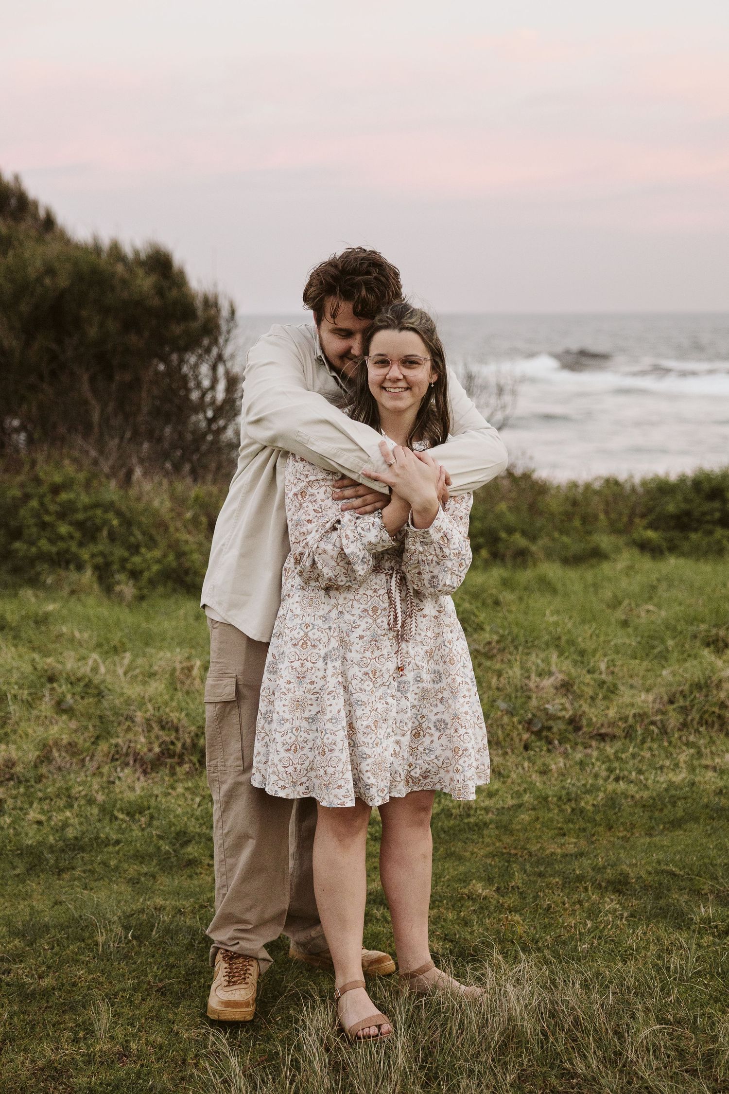 Man embracing woman from behind on grassy coastal cliff at sunset, both smiling toward camera.