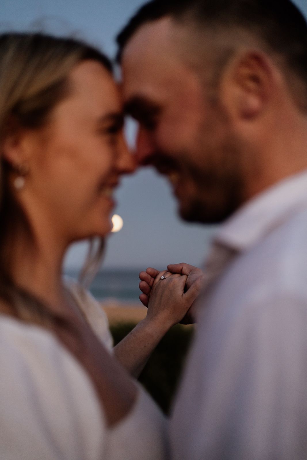 Engaged couple touching foreheads with focus on clasped hands showing diamond ring at sunset.