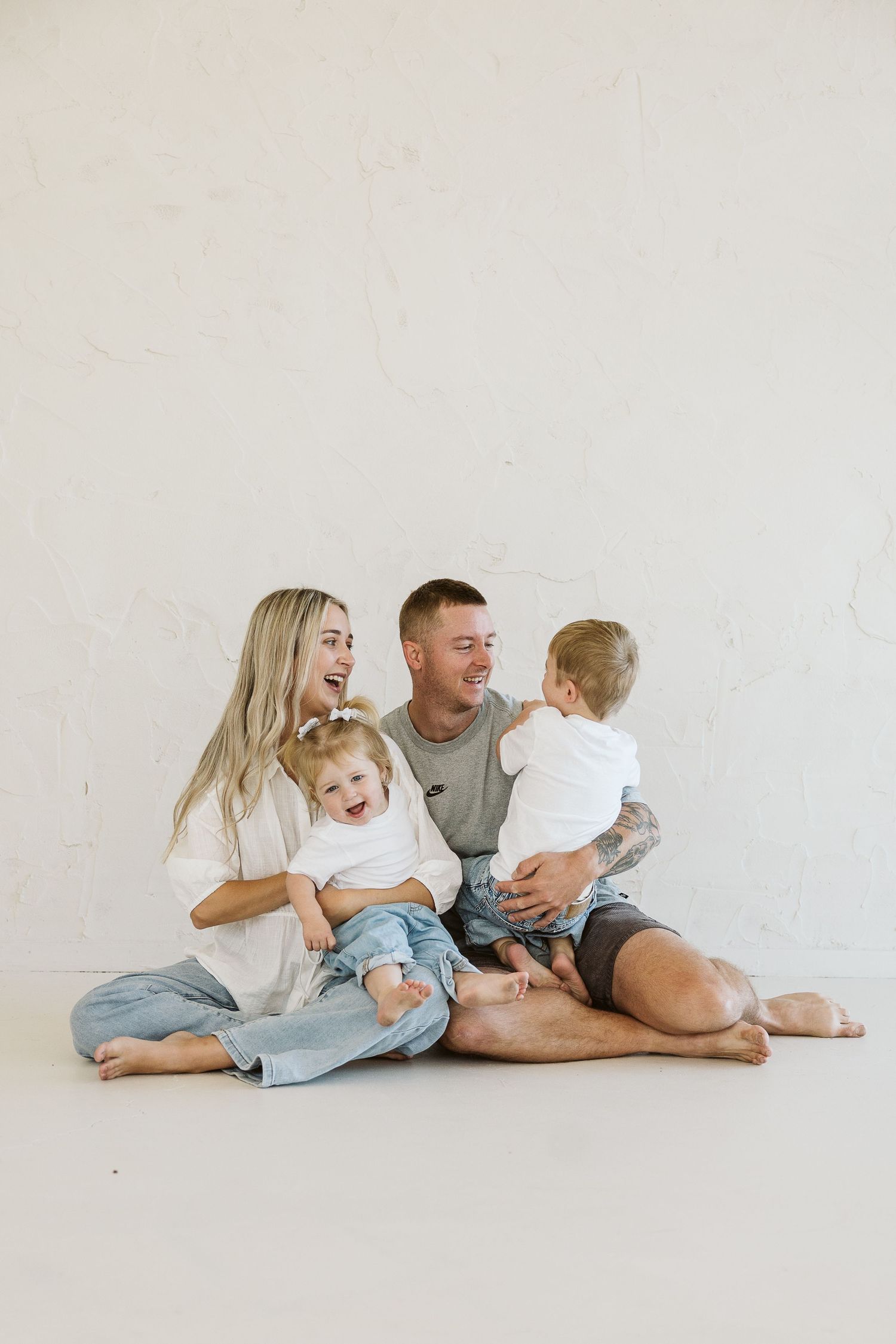 Happy young family with two toddlers sitting together on floor, laughing and bonding in bright white studio.