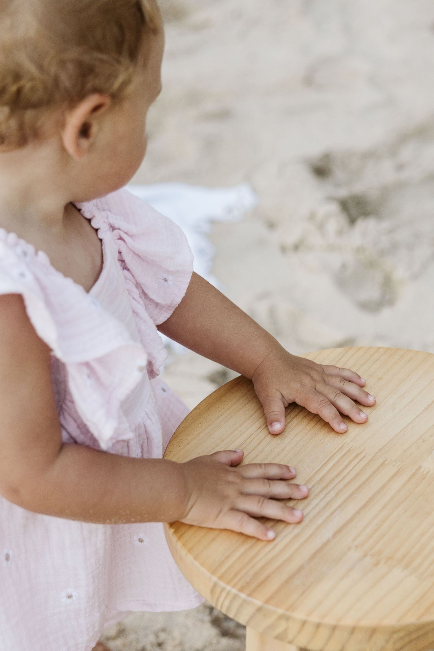 A toddler girl in a pink dress places her hands on a small wooden table while playing on a sandy beach.