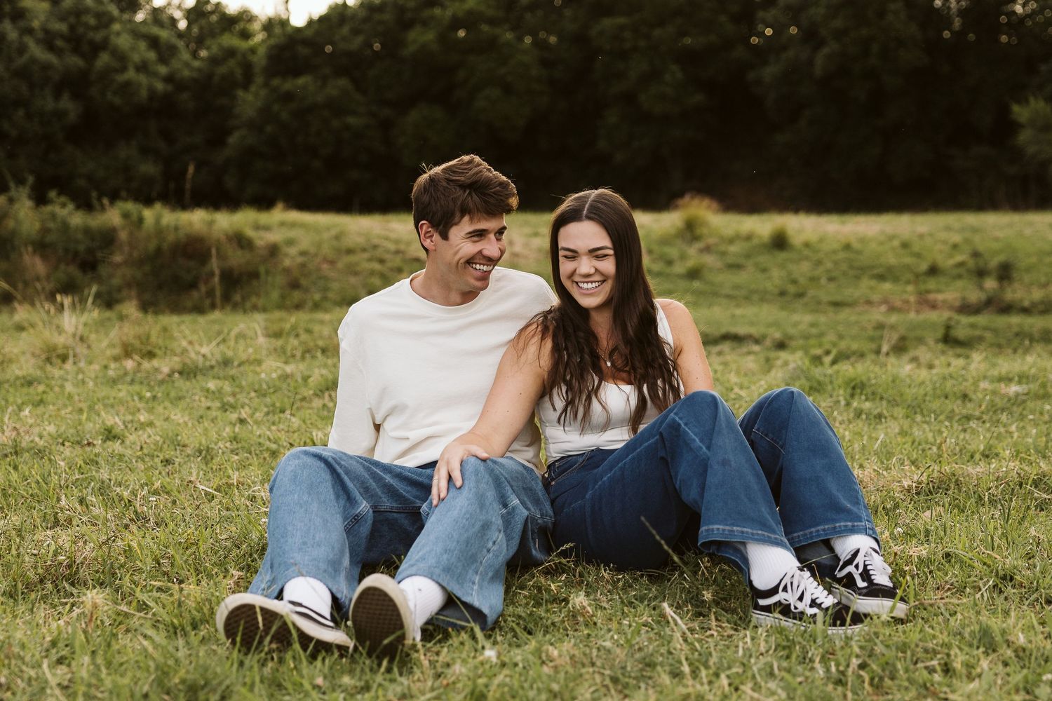 Young smiling couple sitting together on green grass field with trees in background.