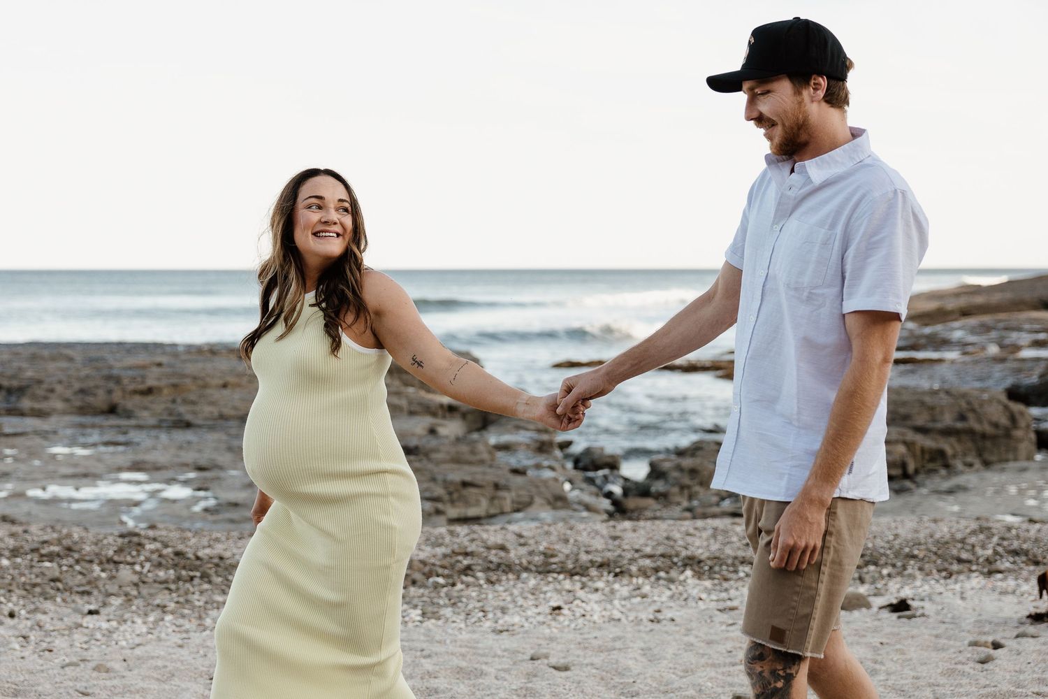 Joyful pregnant couple holds hands smiling on rocky beach with ocean backdrop.
