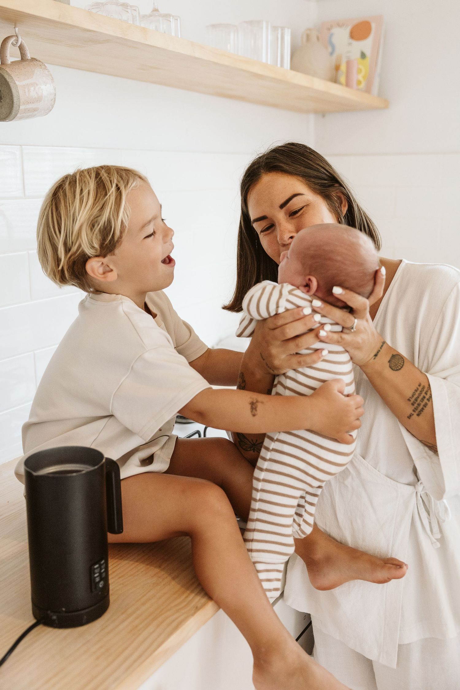 Mother kissing newborn baby while smiling toddler reaches out in a bright white kitchen.