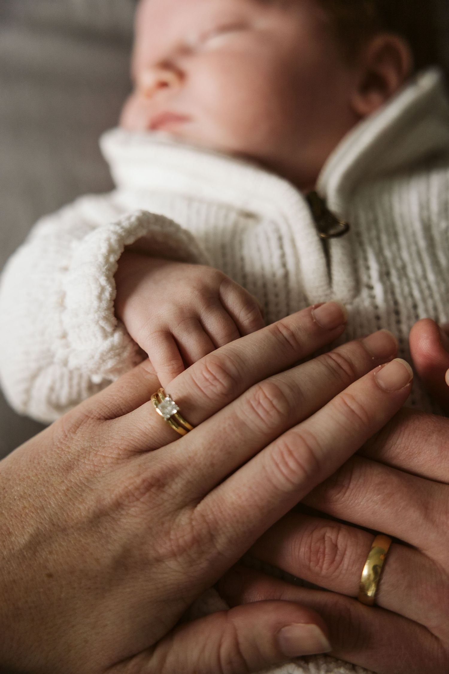 Married couple's hands with wedding rings resting on sleeping newborn baby.