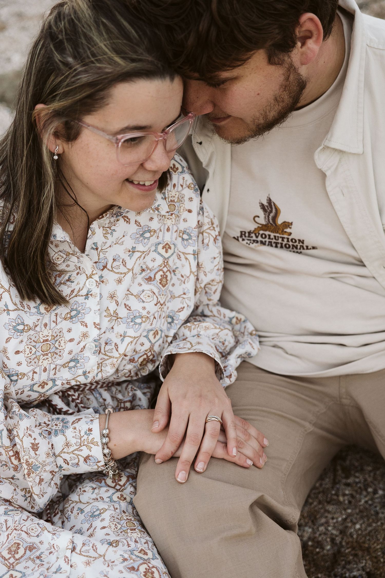 Couple sitting closely together outdoors, woman in floral dress showing engagement ring while smiling.