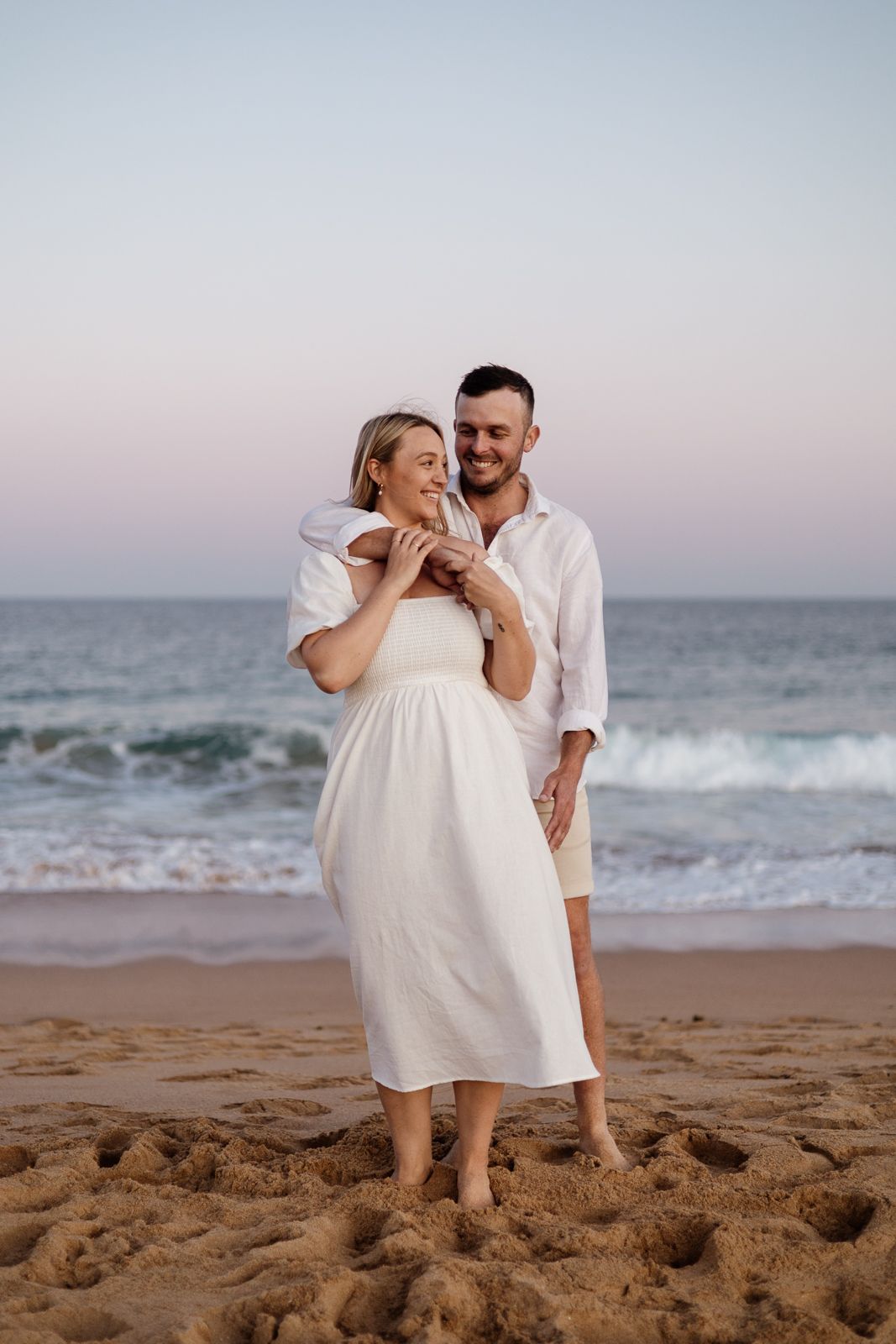 Couple embracing on sandy beach at dusk, woman in white off-shoulder dress, ocean waves behind.