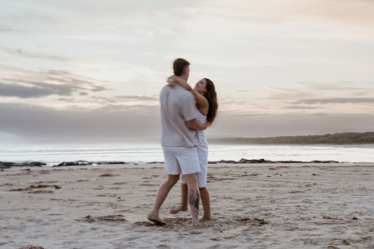 Romantic couple hugging on a sandy beach at sunset, both dressed in white casual clothing with gentle waves behind them.