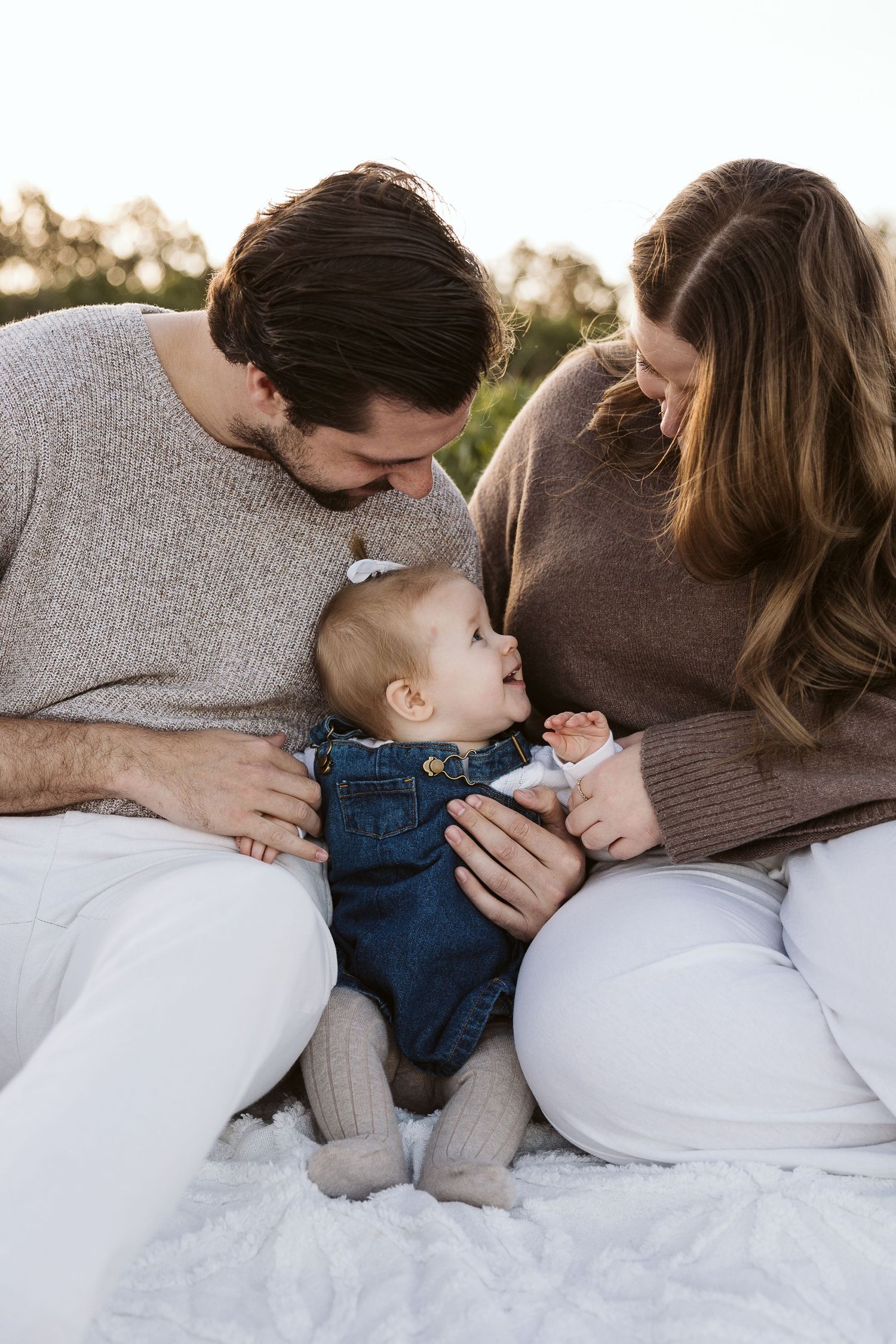 A young family sits outdoors on a white blanket, parents lovingly leaning toward their baby girl in denim overalls.