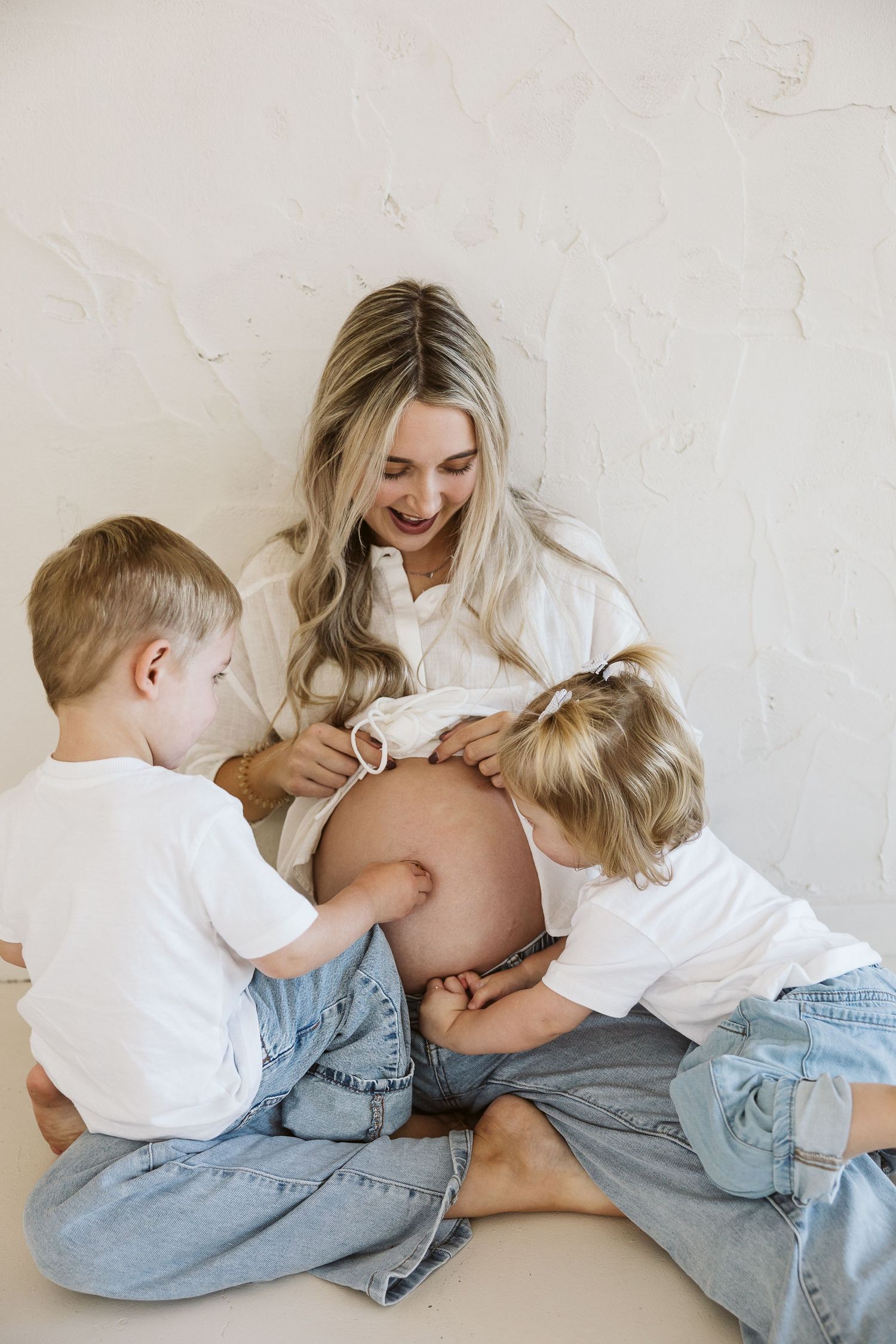 Pregnant woman smiling as two young blonde children touch her baby bump during a maternity photo session.