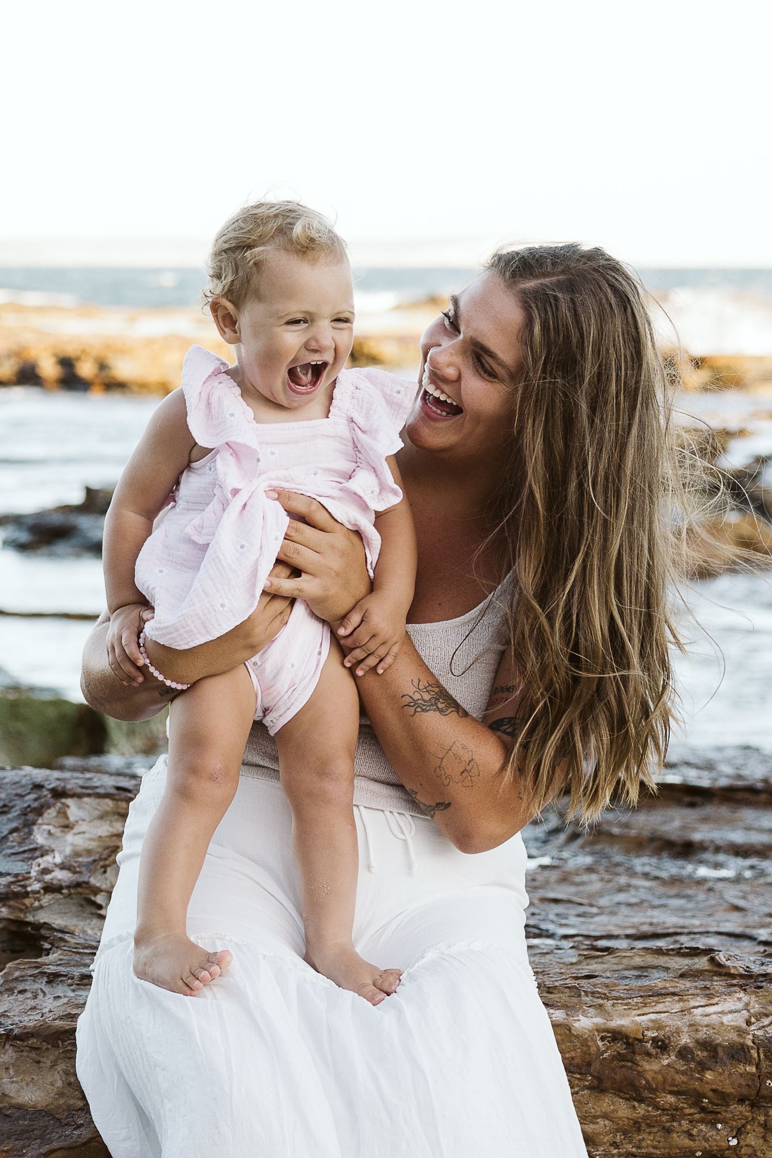 A laughing mother in a white dress holds her giggling baby girl in a pink outfit on a rocky beach.