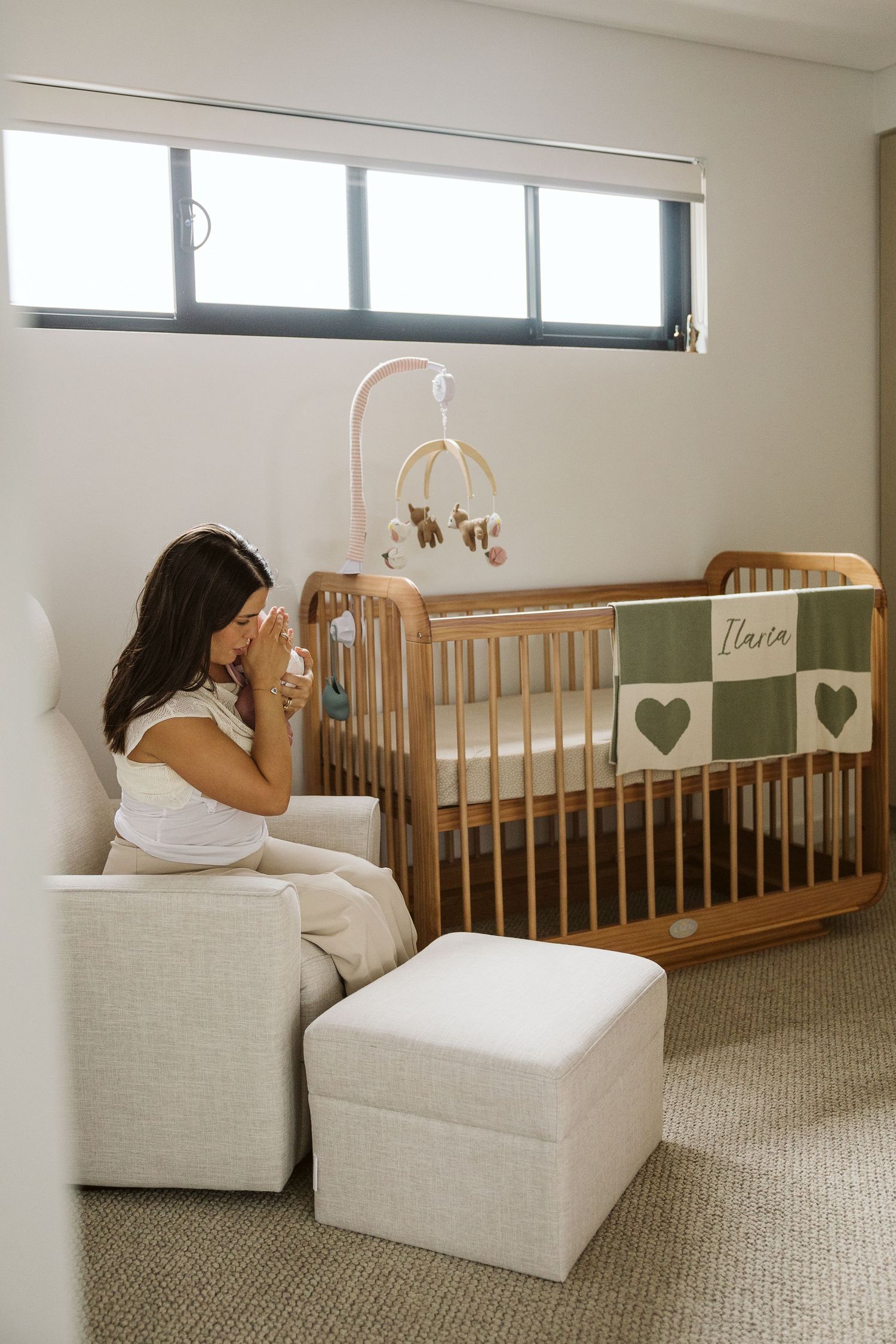 Mother sitting in nursery chair with newborn beside a wooden crib with green heart blanket.