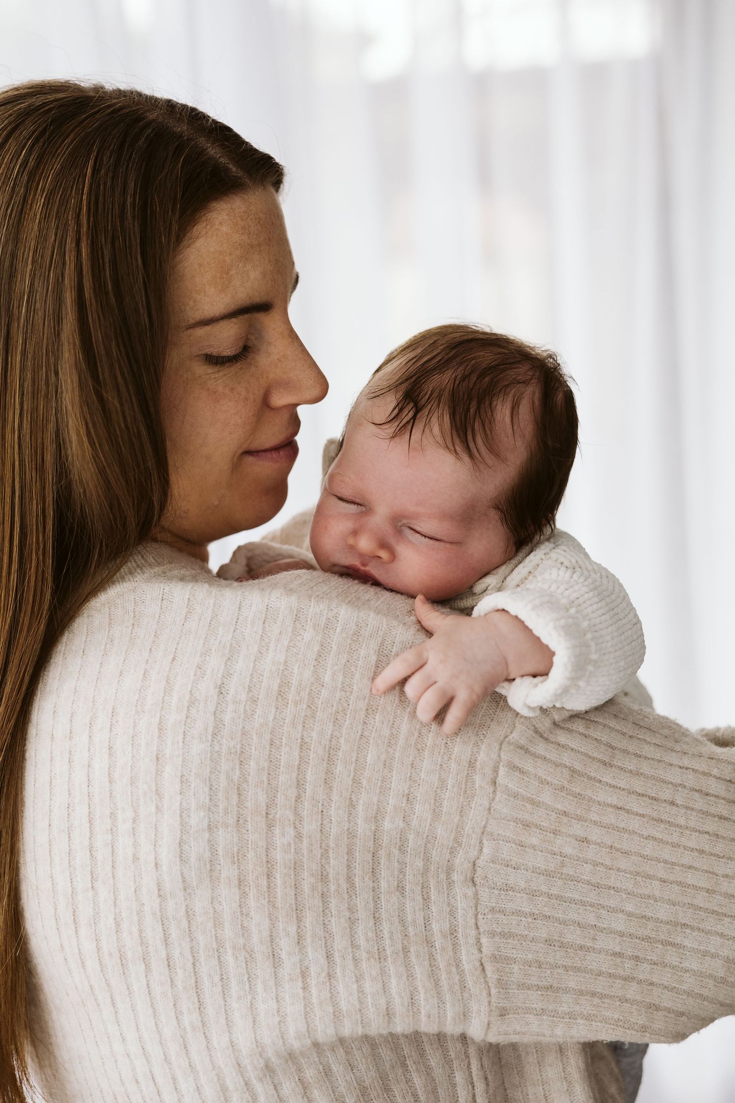 Mother in cream sweater gently holding sleeping newborn on her shoulder.