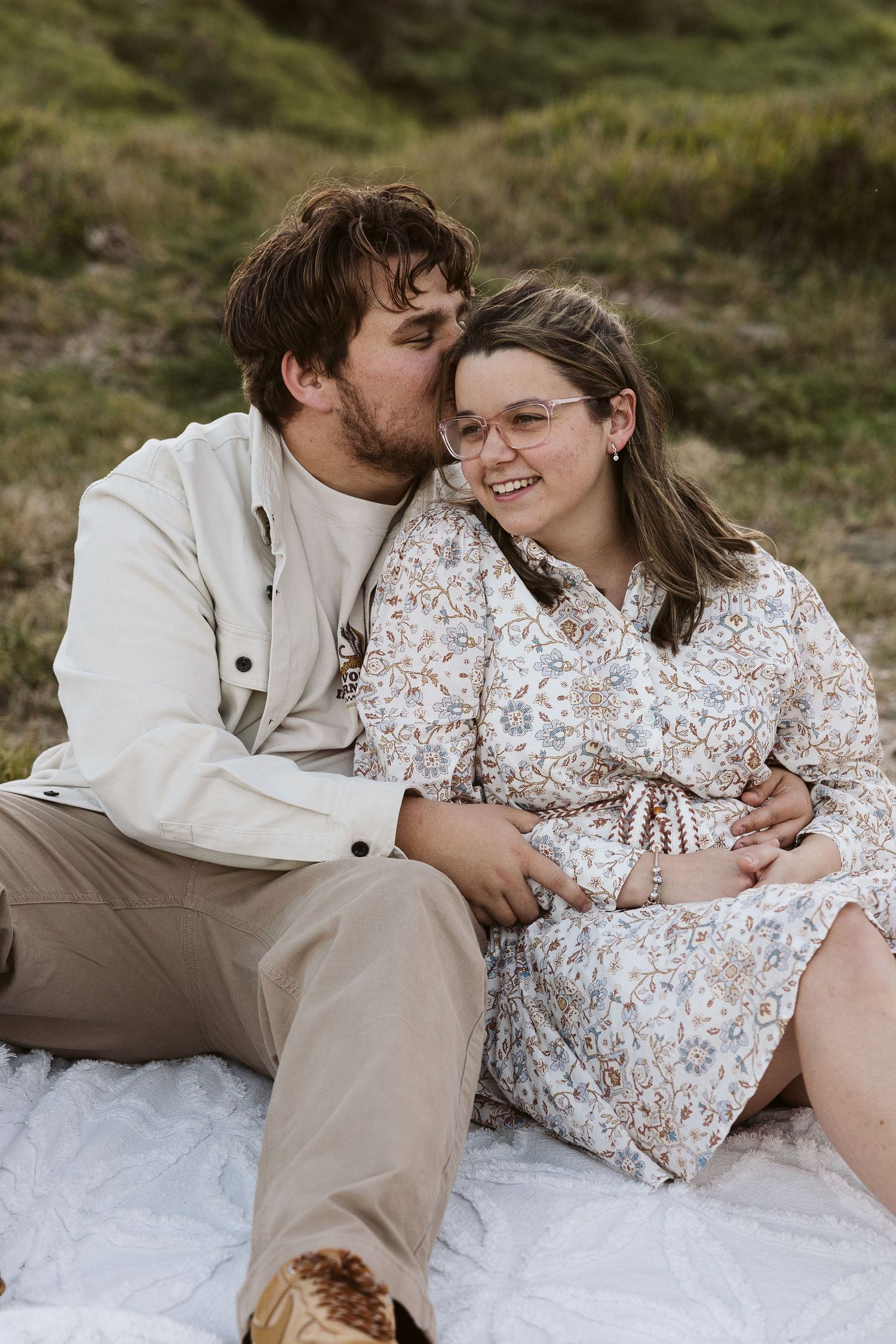 Happy couple sitting on white blanket in green field, man kissing womans cheek as she laughs.