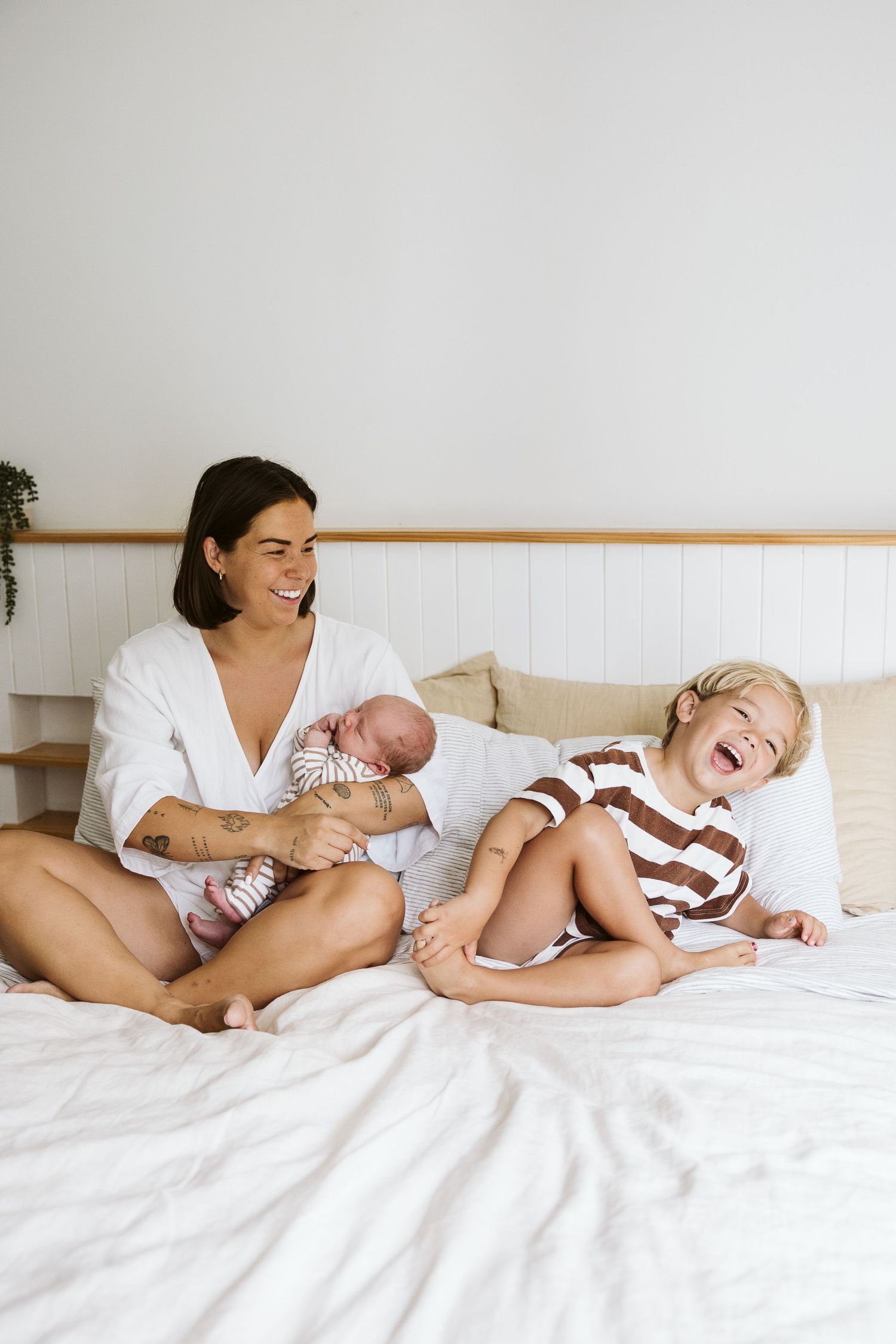 Mother in white robe laughing with newborn and toddler son on a white bed.