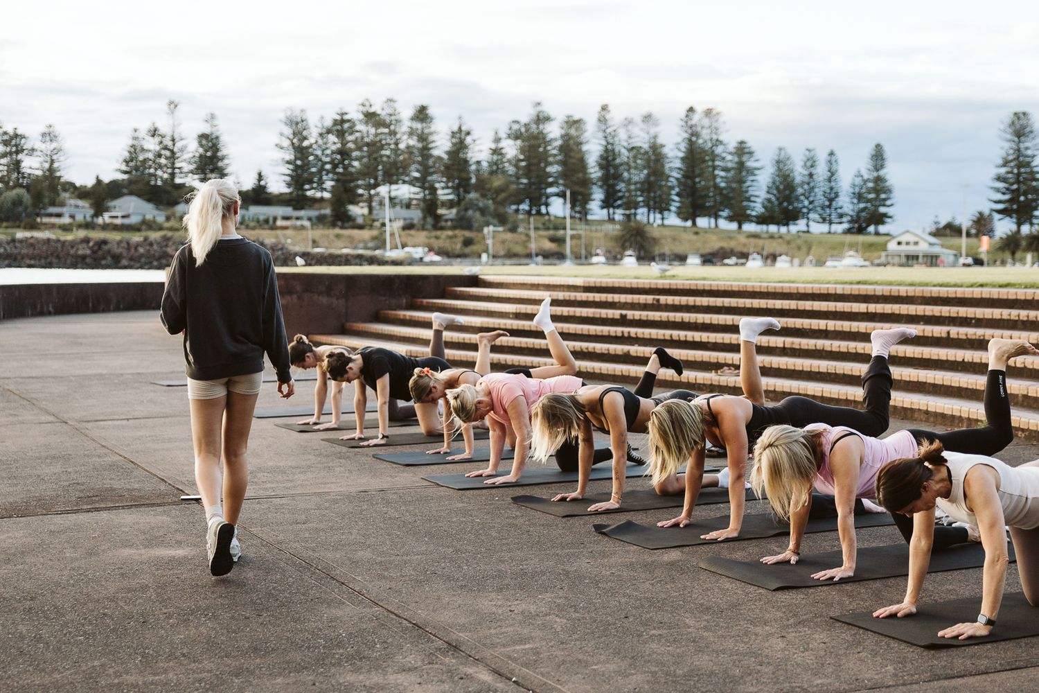 Fitness instructor leading an outdoor group exercise class in downward dog pose near the ocean.