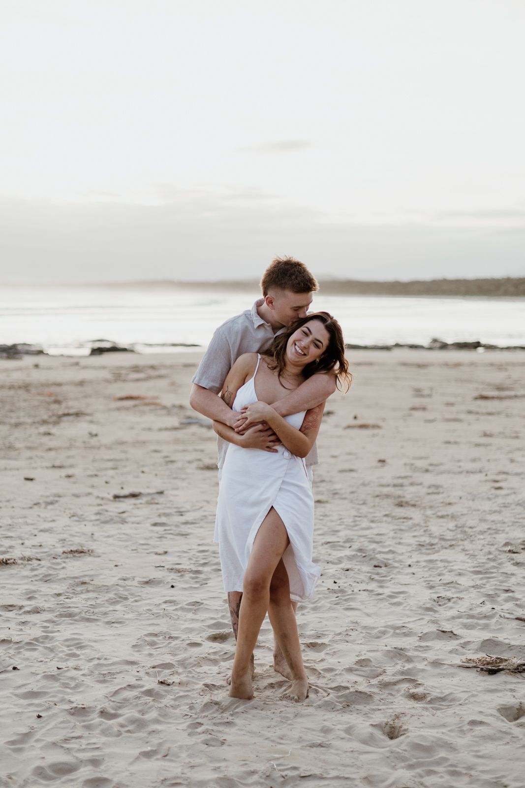 Man hugging woman from behind as she laughs joyfully during a romantic beach couples photo session.