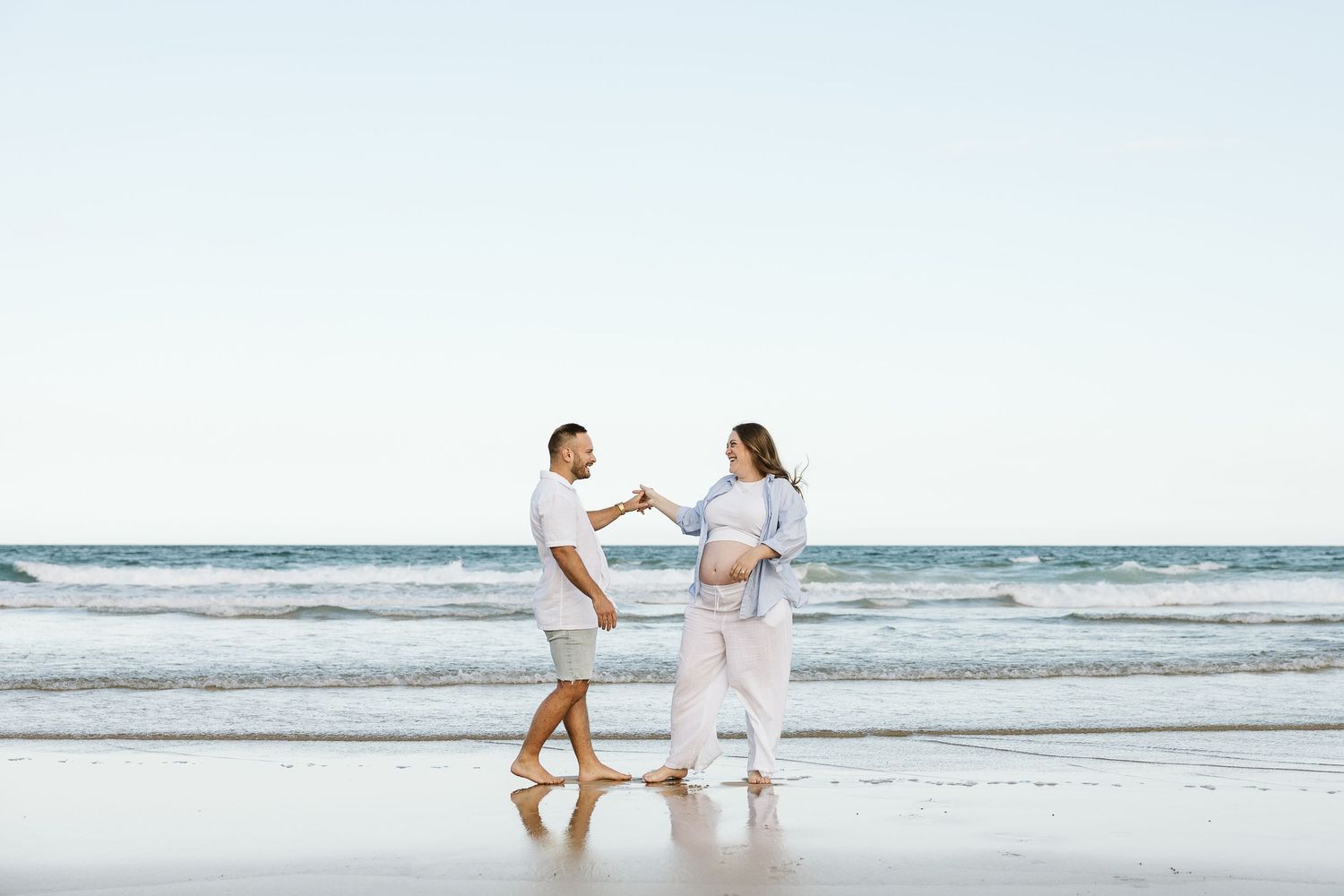Expecting couple holding hands and facing each other on reflective wet sand beach with ocean waves.