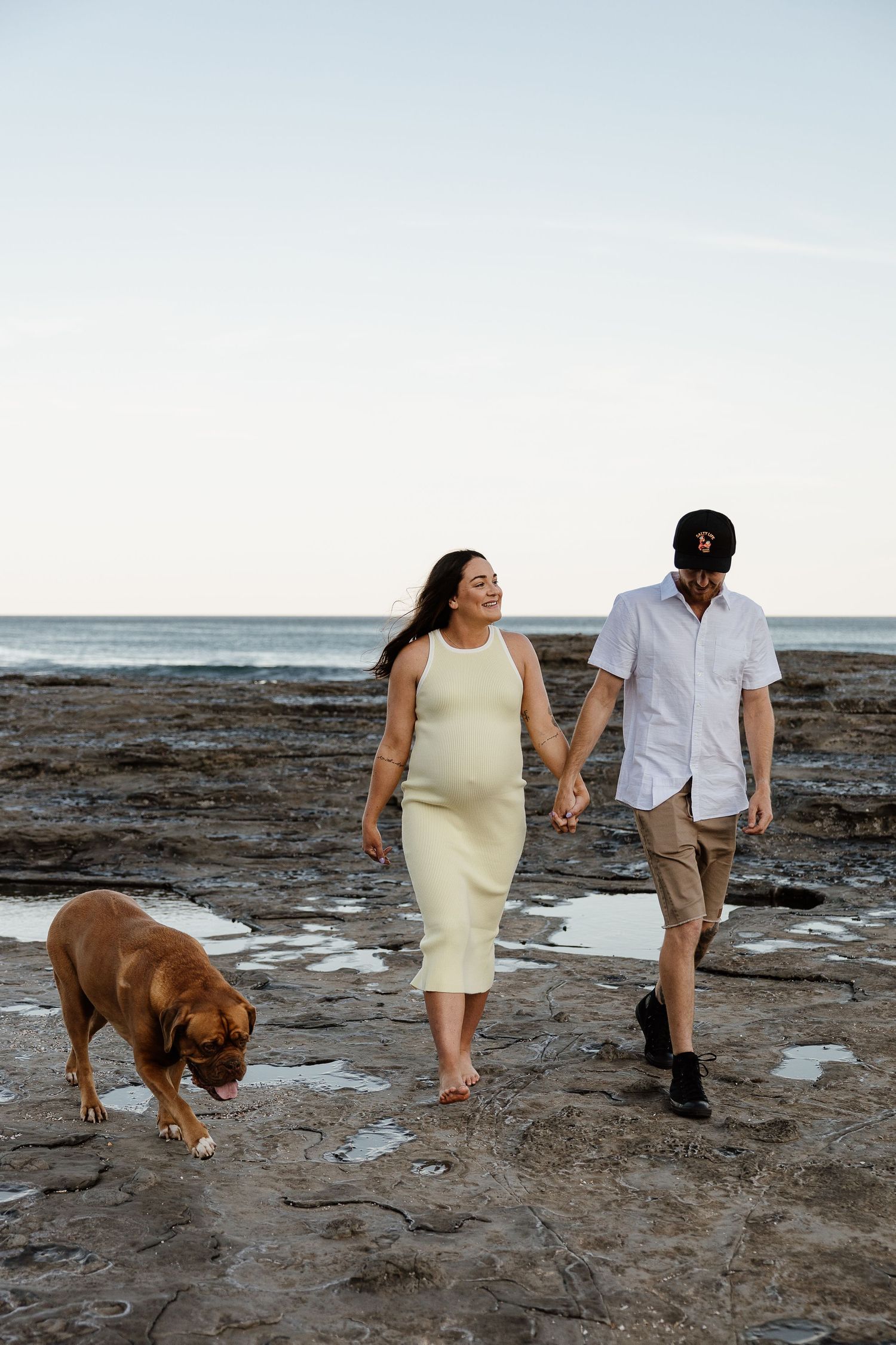 Smiling pregnant couple holds hands walking on rocks with brown dog by ocean.