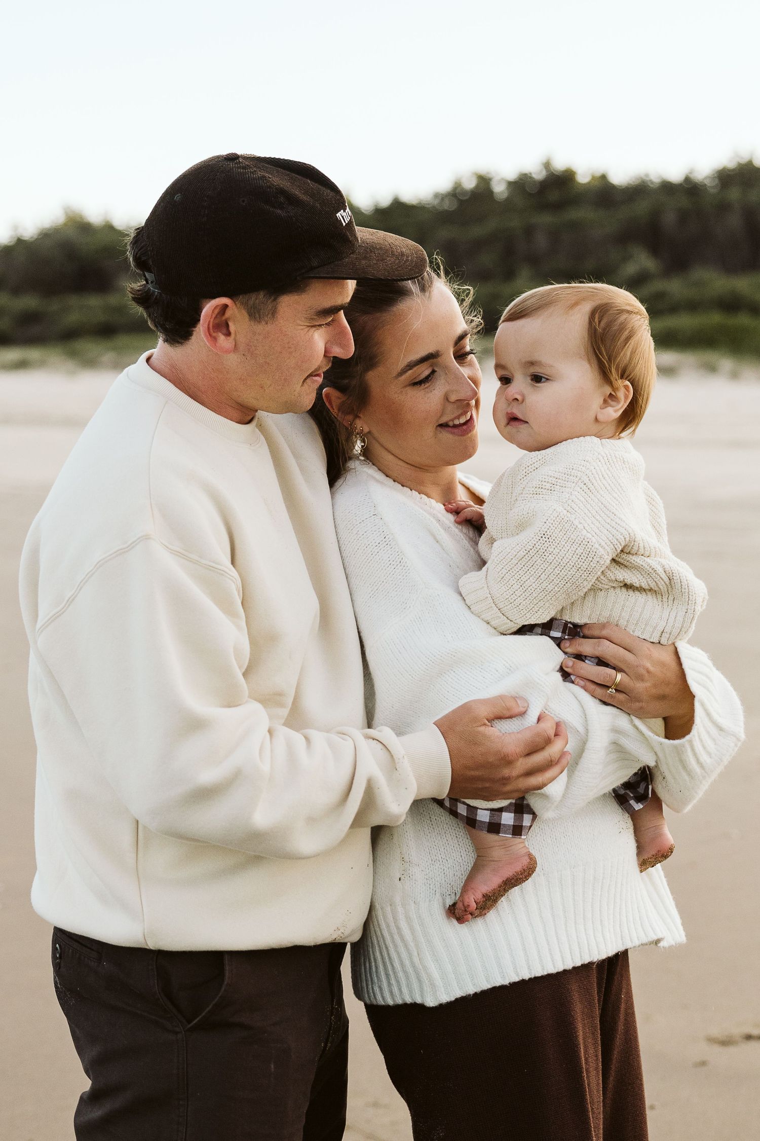 A couple in white sweaters smiles at their baby on a sandy beach with trees in the background.