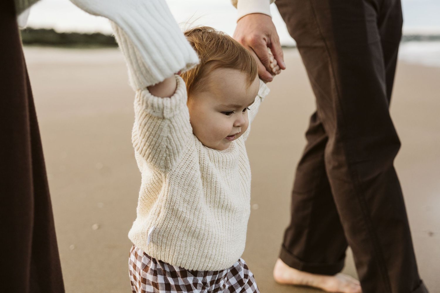 Toddler in white knit sweater holds parents hands while learning to walk on sandy beach.