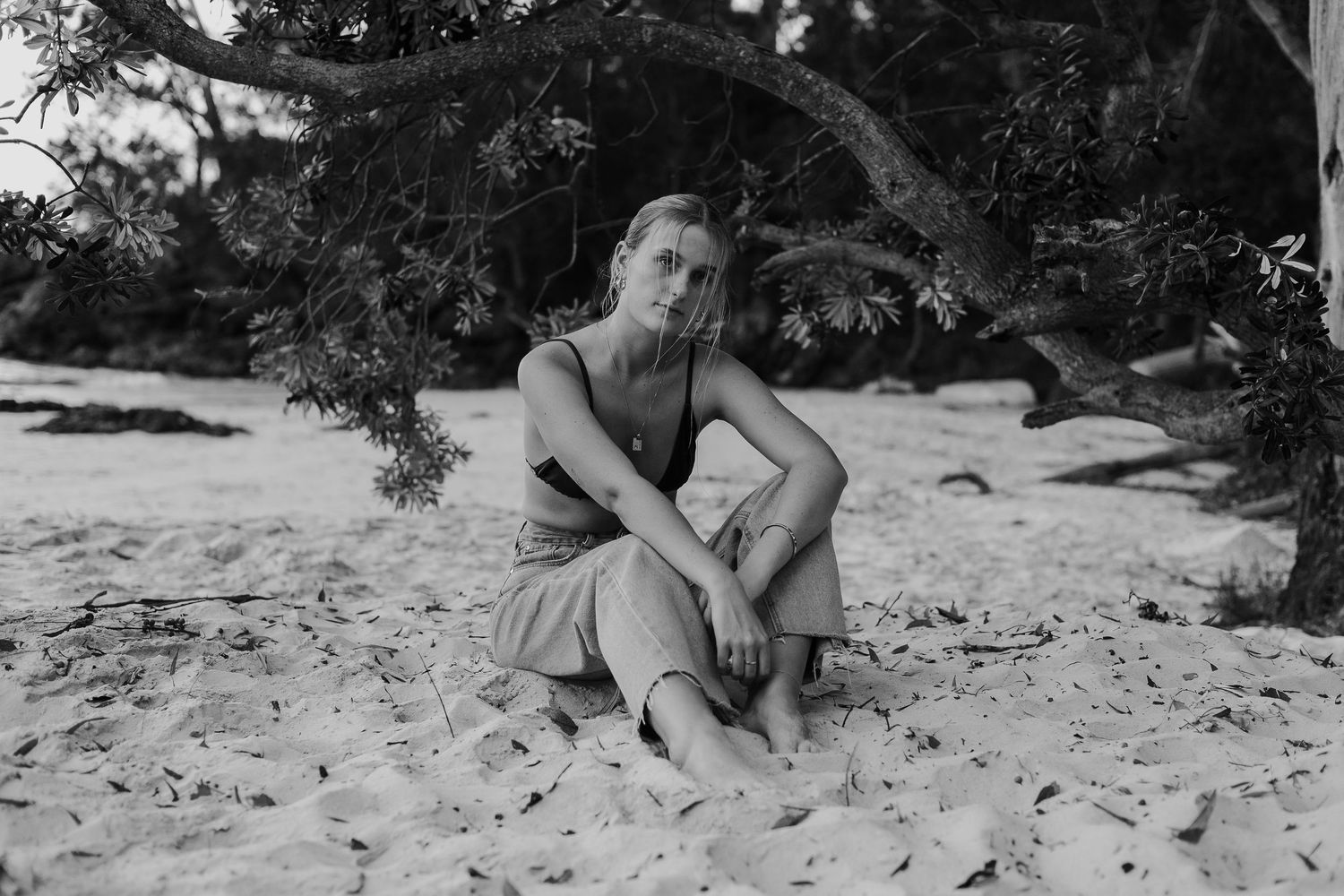 Black and white portrait of a woman sitting barefoot on sand under trees. Portrait photography.