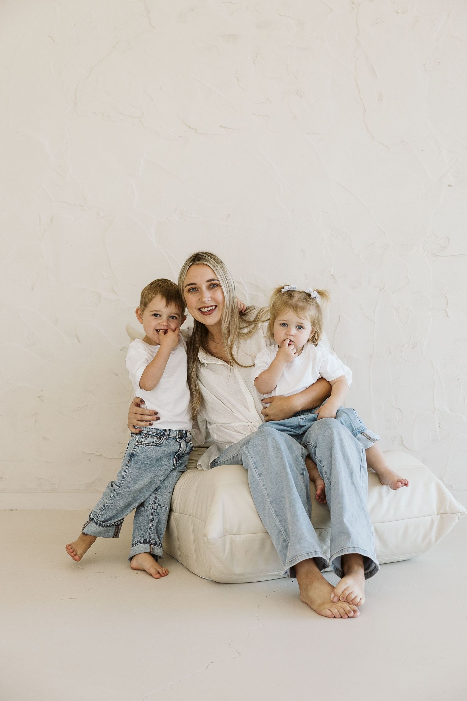 Young woman sitting on white bean bag chair with two toddlers, all wearing jeans and white tops in bright studio.