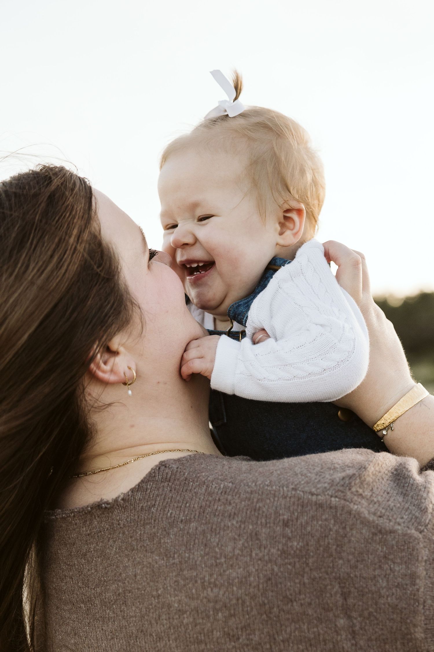 A joyful baby laughing while being kissed on the cheek by their mother outdoors in warm natural light.