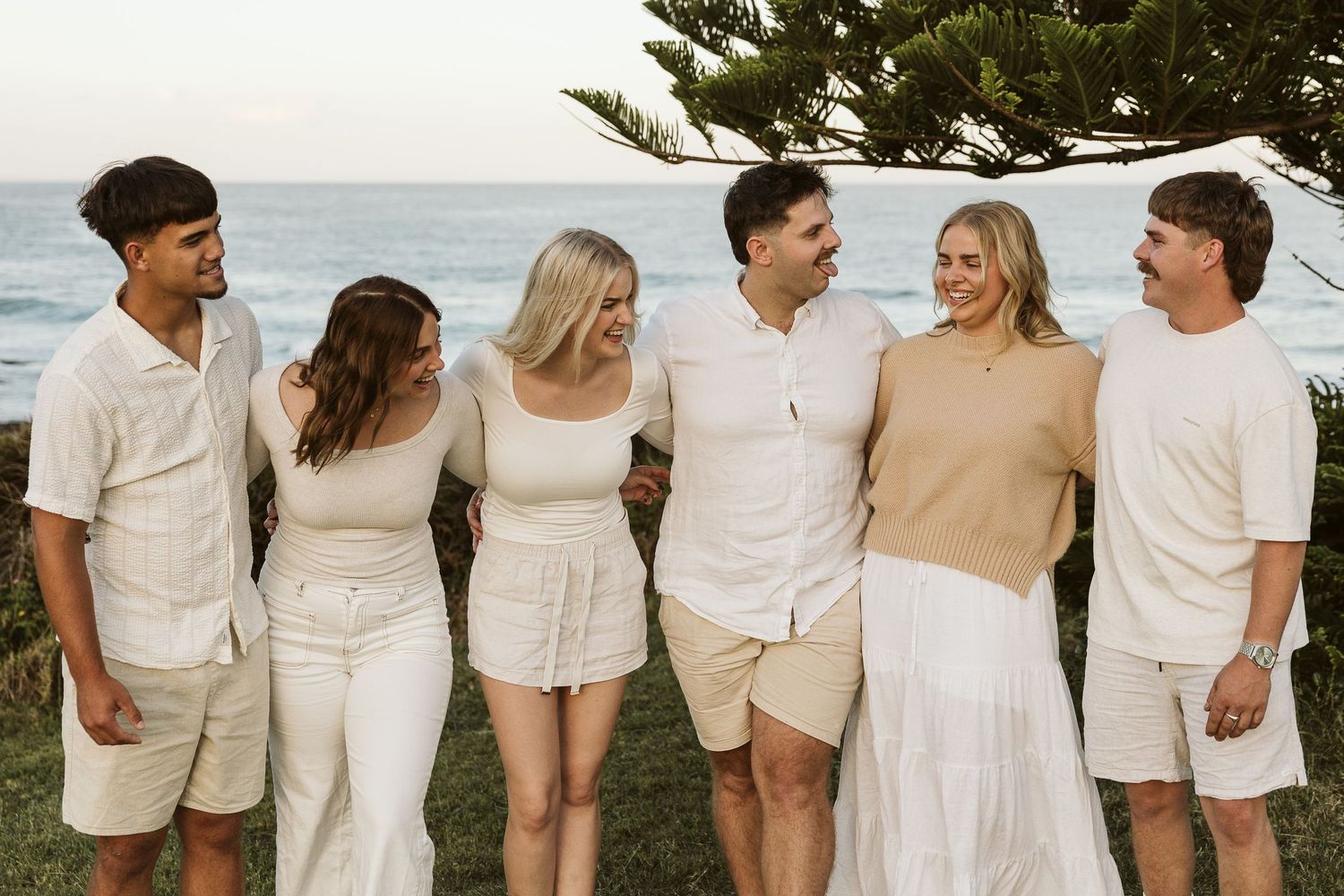 Six friends in coordinating white and beige outfits laughing together on a grassy coastal overlook with ocean views.