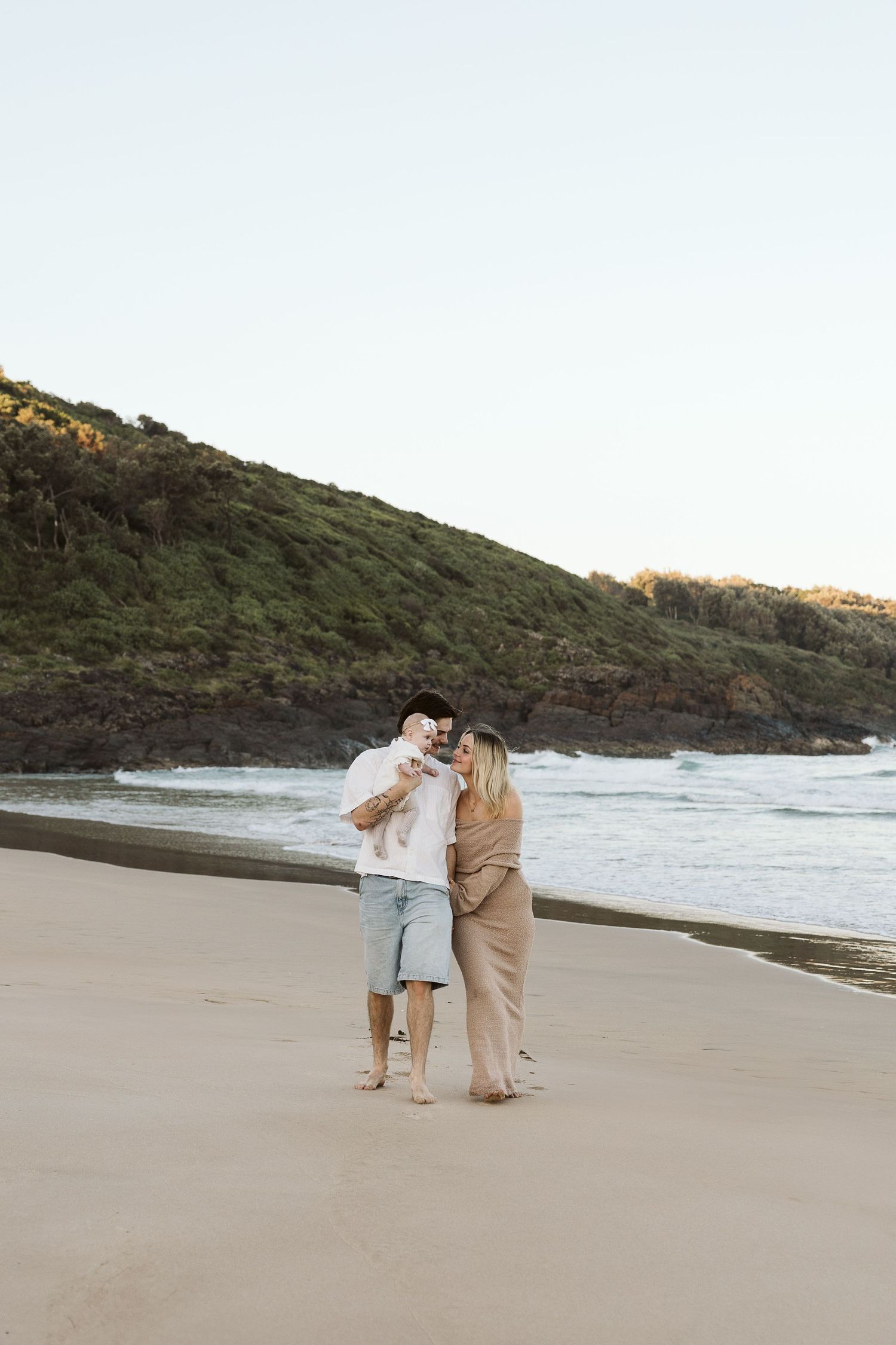Happy couple holding baby on scenic beach with green hillside and ocean waves in background.