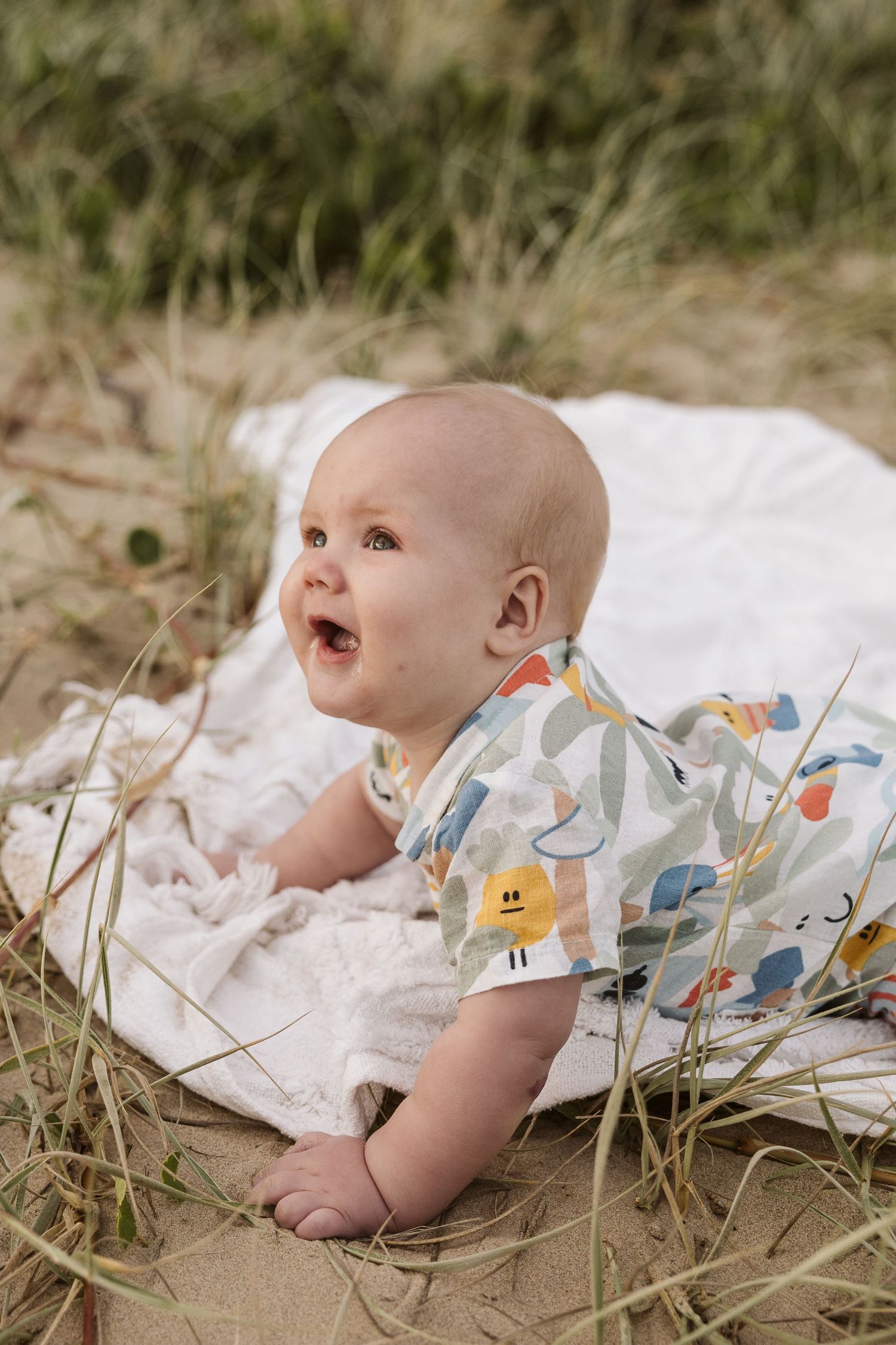 A happy baby in a colorful printed onesie lying on a white blanket outdoors on sandy ground with grass.