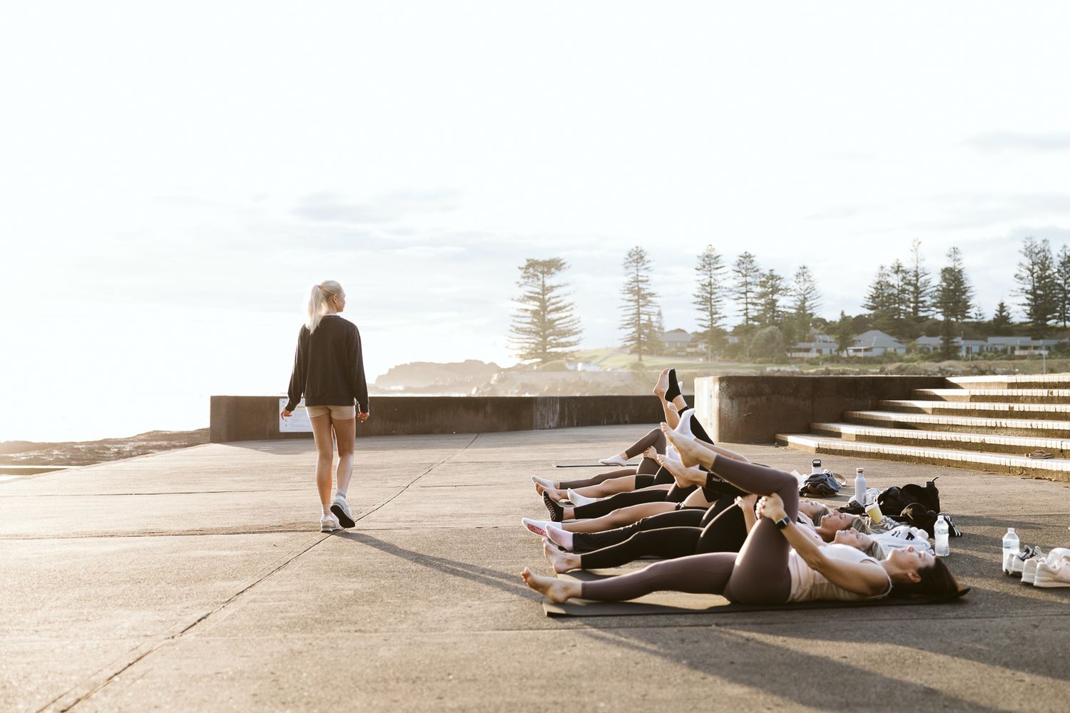Outdoor fitness instructor walks past group exercising on mats at scenic beachside location.