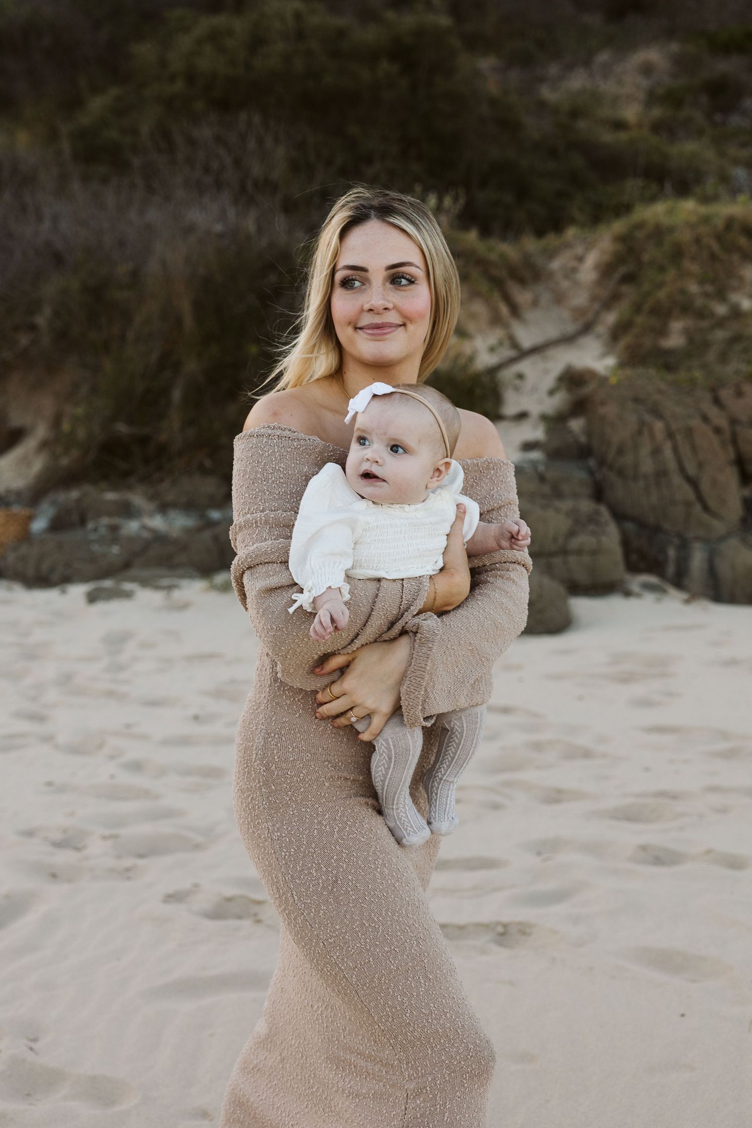 A blonde woman in a tan knit dress holds her baby girl on a sandy beach.