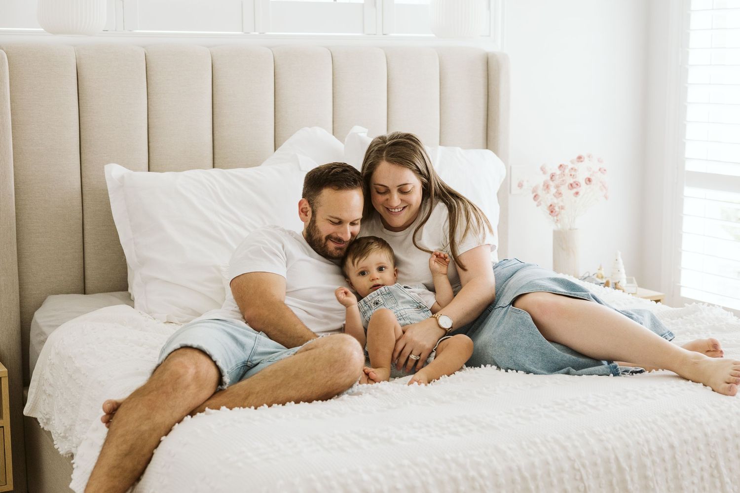 A happy young family with a baby relaxing together on a white bed in a bright, airy bedroom.