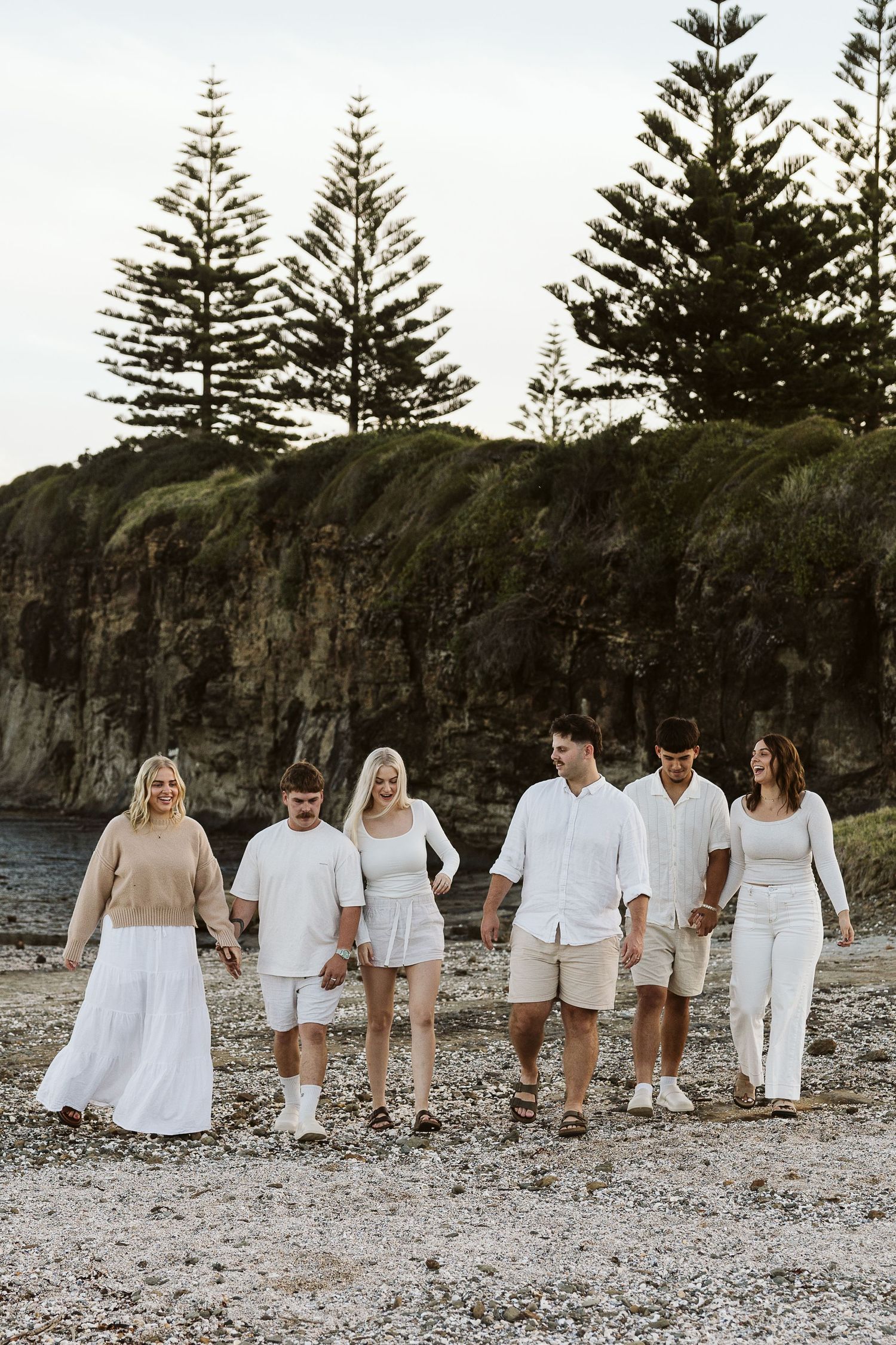 Group of six people in white and beige outfits walking together on a rocky beach with green cliffs behind.