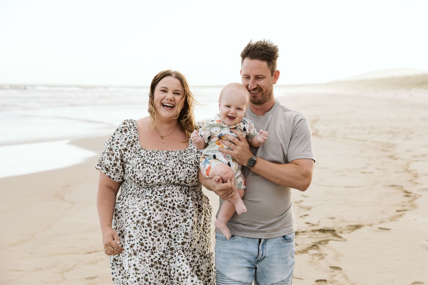 Happy family with baby strolling on sandy beach, parents smiling, casual summer attire, ocean waves in background.