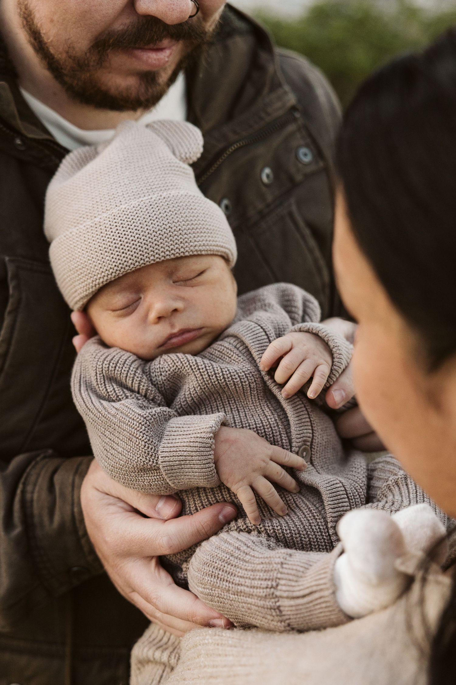 Sleeping newborn baby in beige knit hat and outfit being held gently by both parents outdoors.